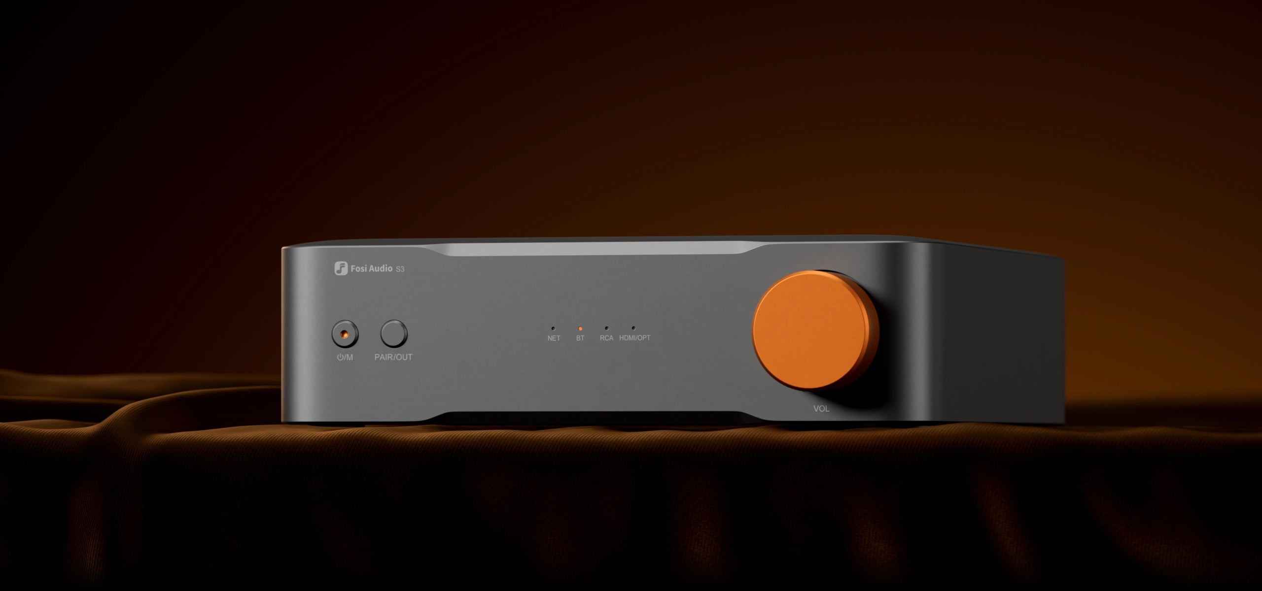 Grijze Fosi Audio S3 versterker met oranje volumeknop op donkere stof.