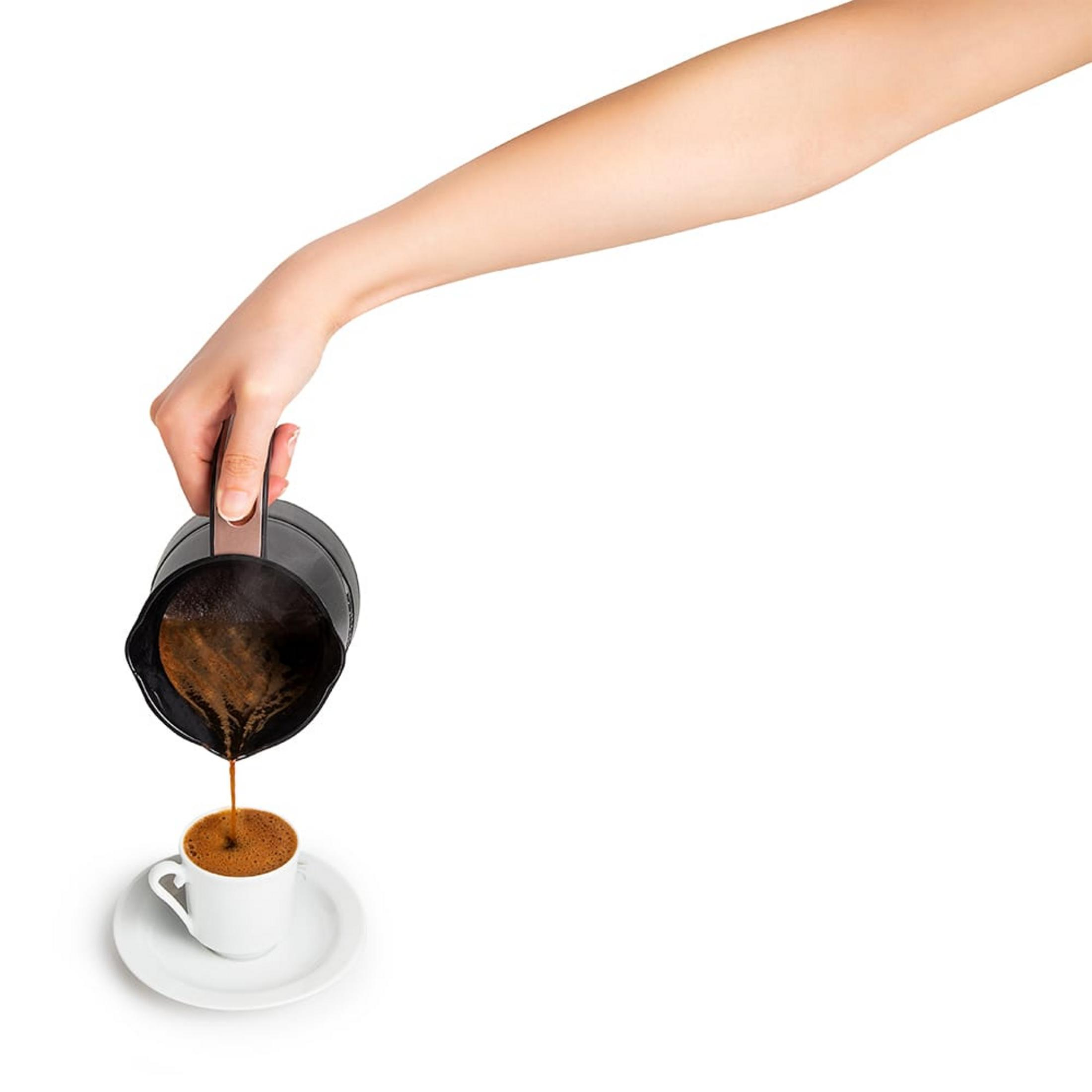 Eine Hand gießt dunklen Kaffee aus einem schwarzen Topf in eine weiße Tasse.