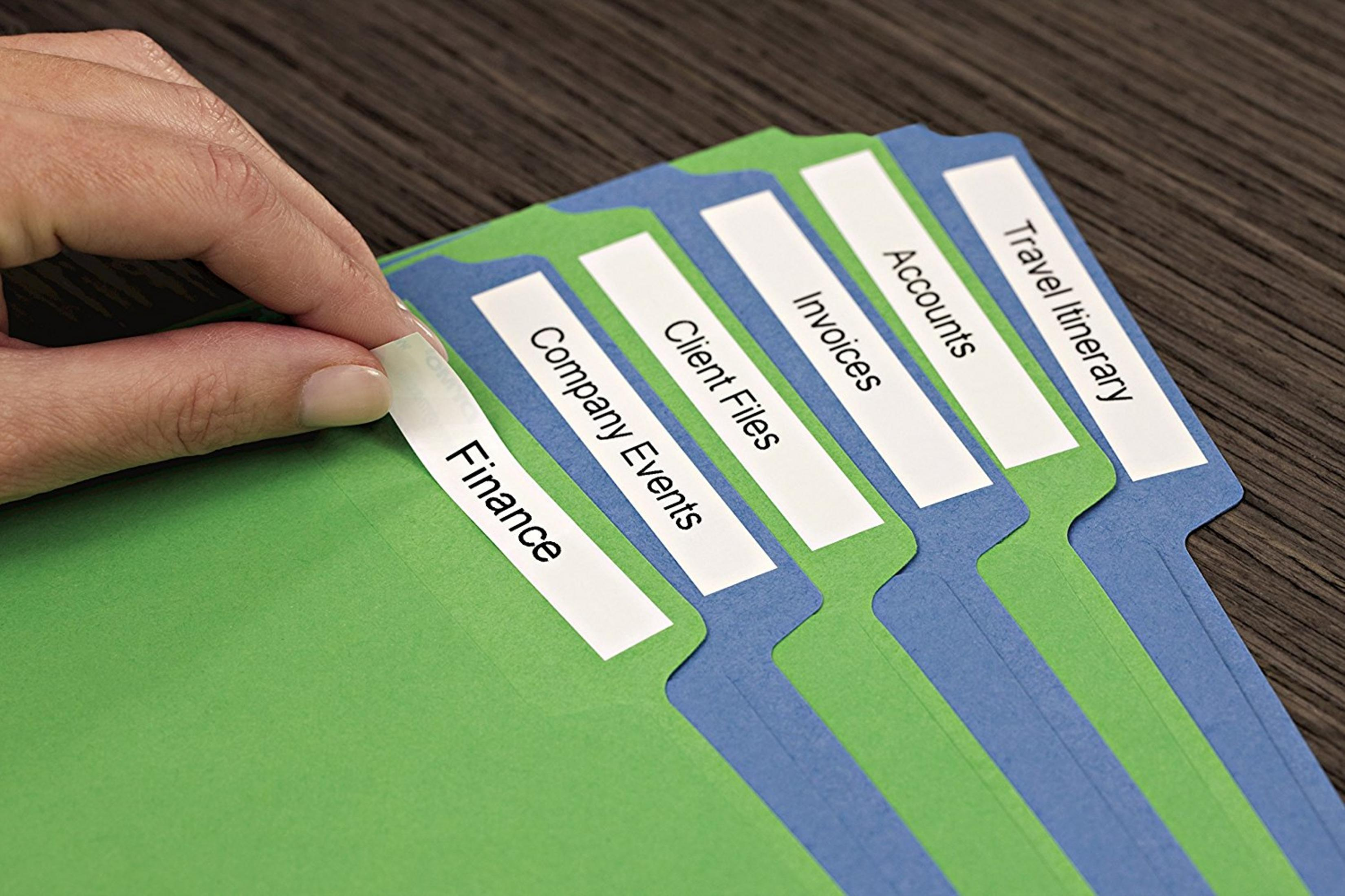 Eine Hand zieht ein Etikett von einer grünen Mappe neben blauen Mappen mit den Aufschriften Finanzen, Firmenveranstaltungen, Rechnungen, Konten, Reiseplan.