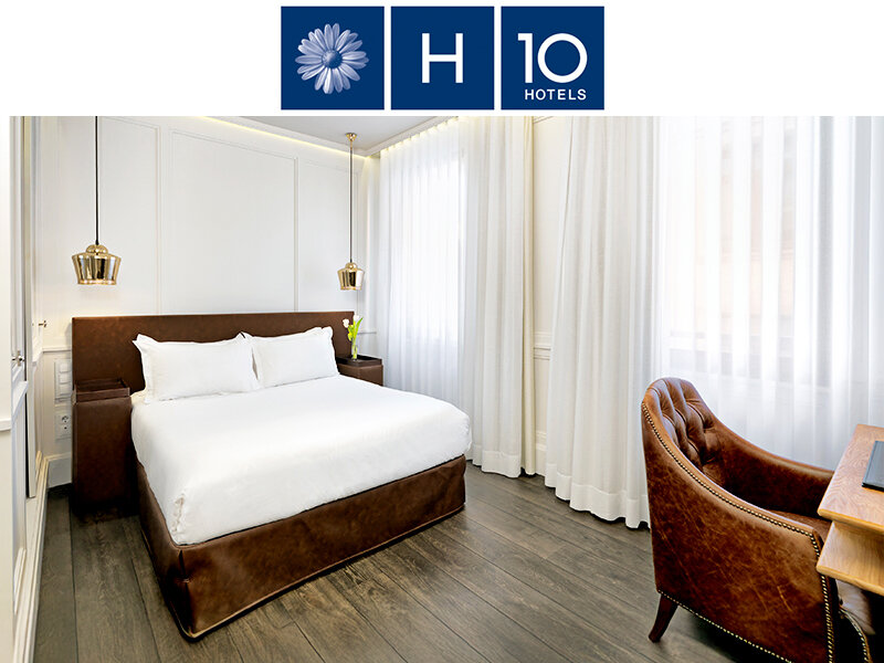 Una habitación de hotel con cama, sillón y escritorio. Paredes blancas, suelo de madera oscura.