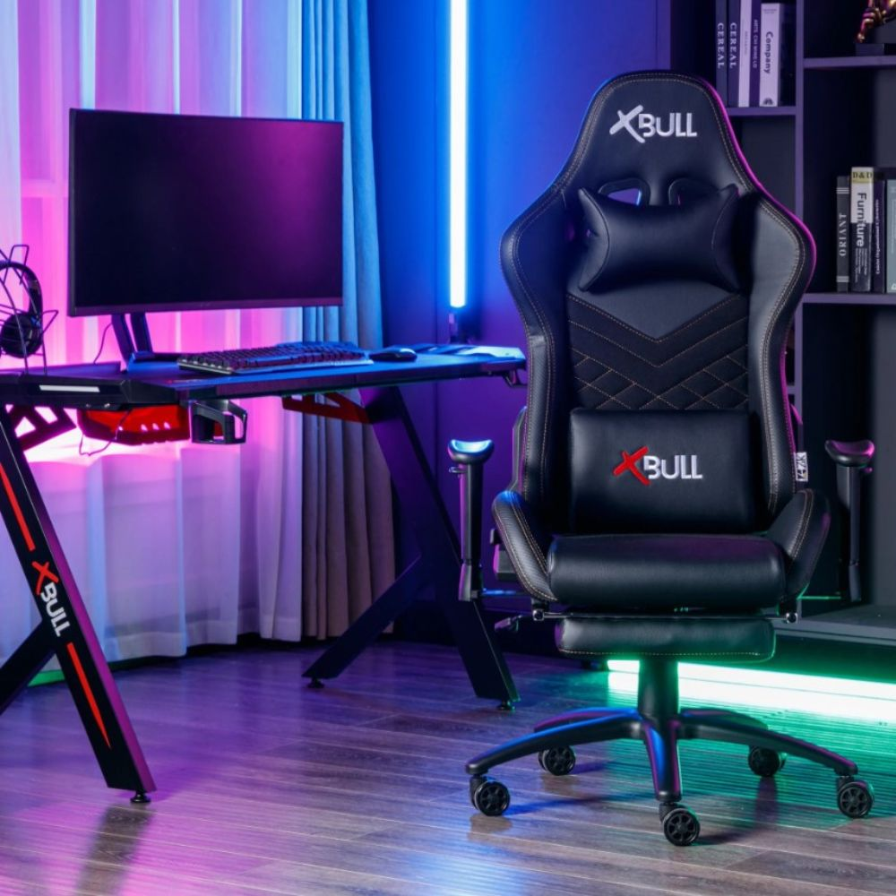 Una sedia da gaming nera con poggiatesta e cuscino lombare siede accanto a una scrivania con monitor.