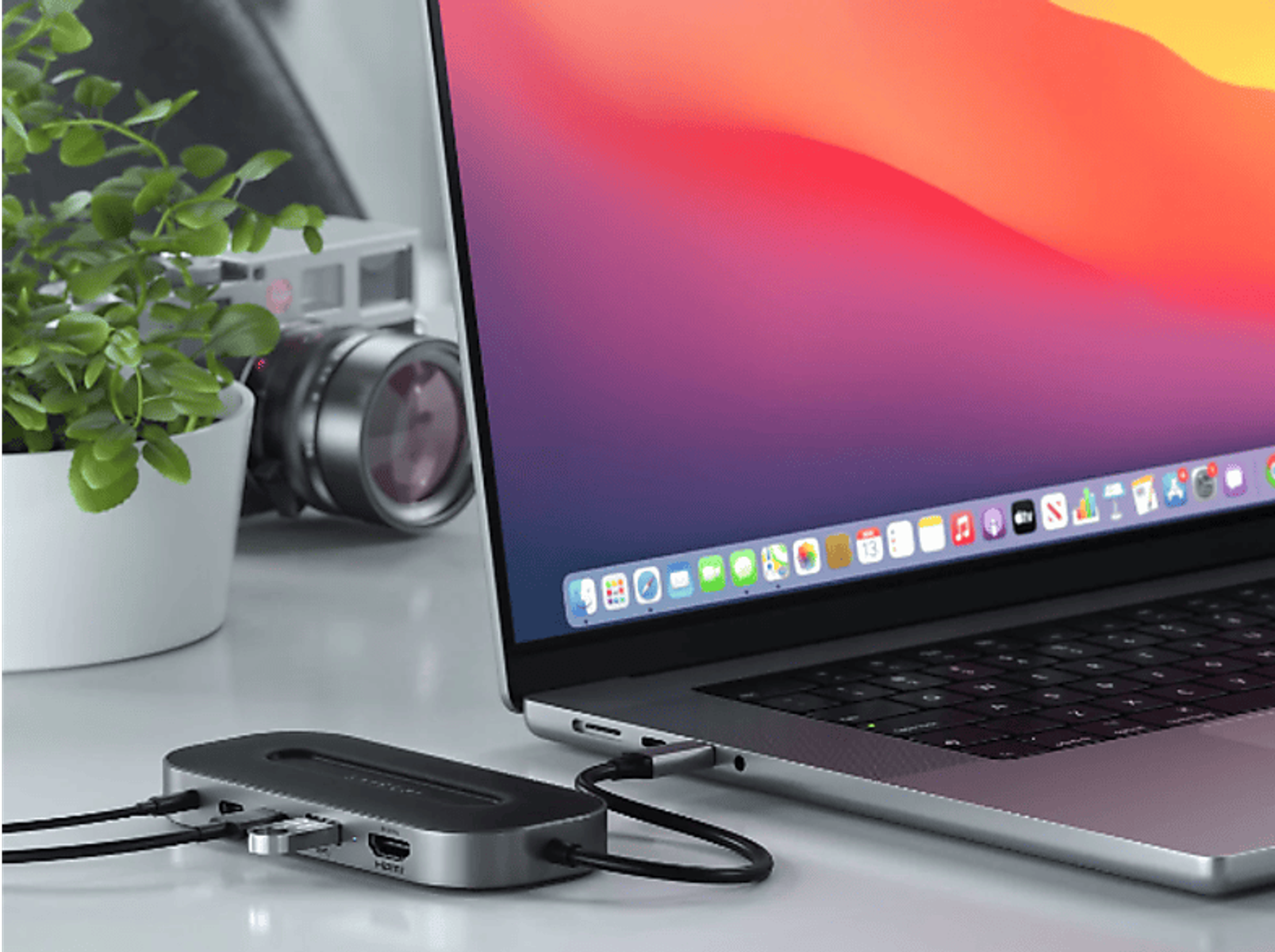 Ein Laptop ist mit einem USB-C-Hub verbunden. Eine Pflanze und eine Kamera sind daneben.