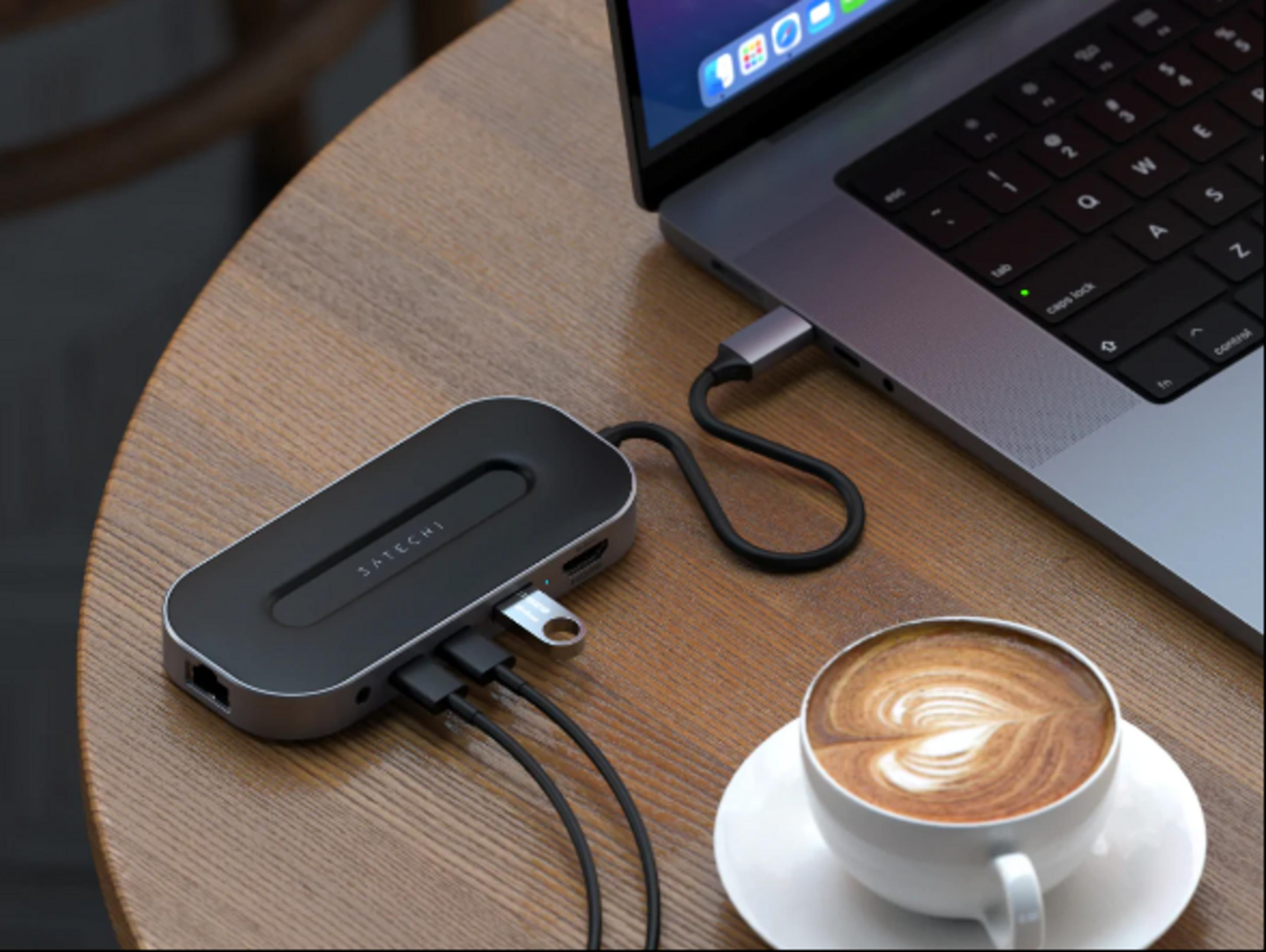 Ein Satechi-Hub ist an einem Laptop angeschlossen, neben einer Kaffeetasse auf einem Holztisch.