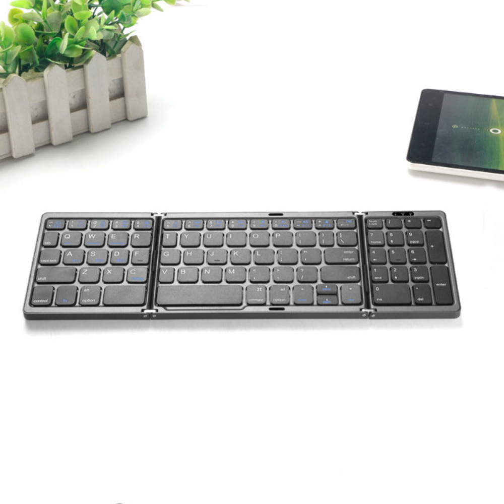 Ein faltbares graues Keyboard liegt auf einem weißen Tisch neben einer Pflanze und einem Tablet.