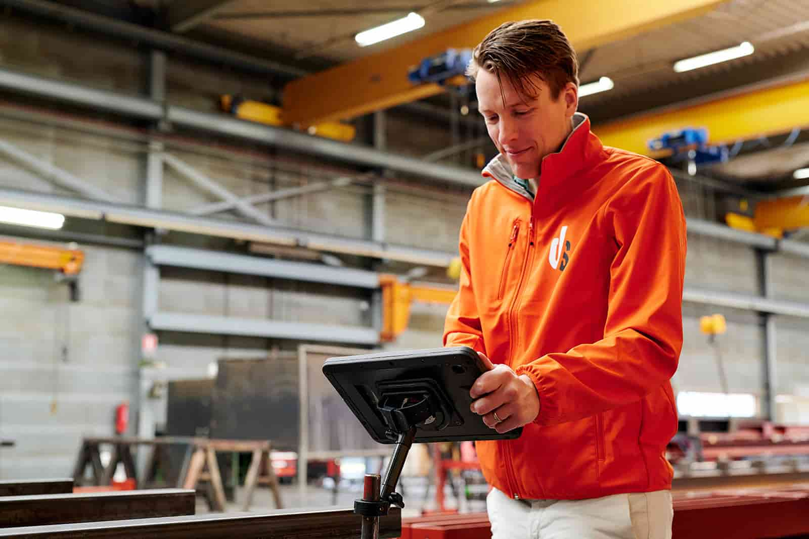 Ein Mann in orangefarbener Jacke bedient ein Tablet in einer Fabrikhalle.