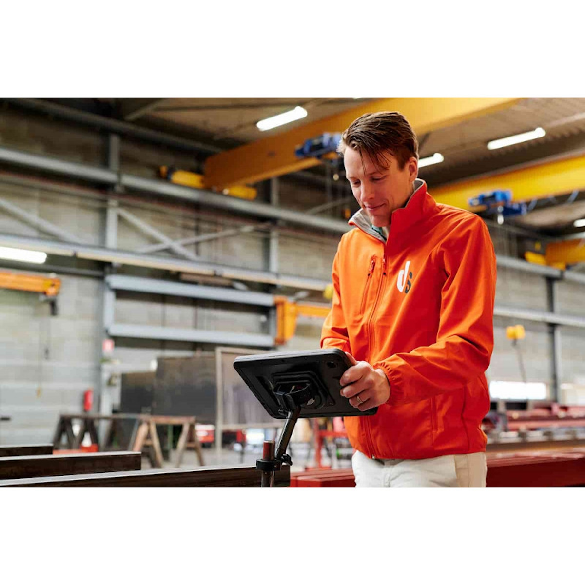 Ein Mann in einer orangefarbenen Jacke steht in einer Fabrikhalle und blickt auf ein Tablet.