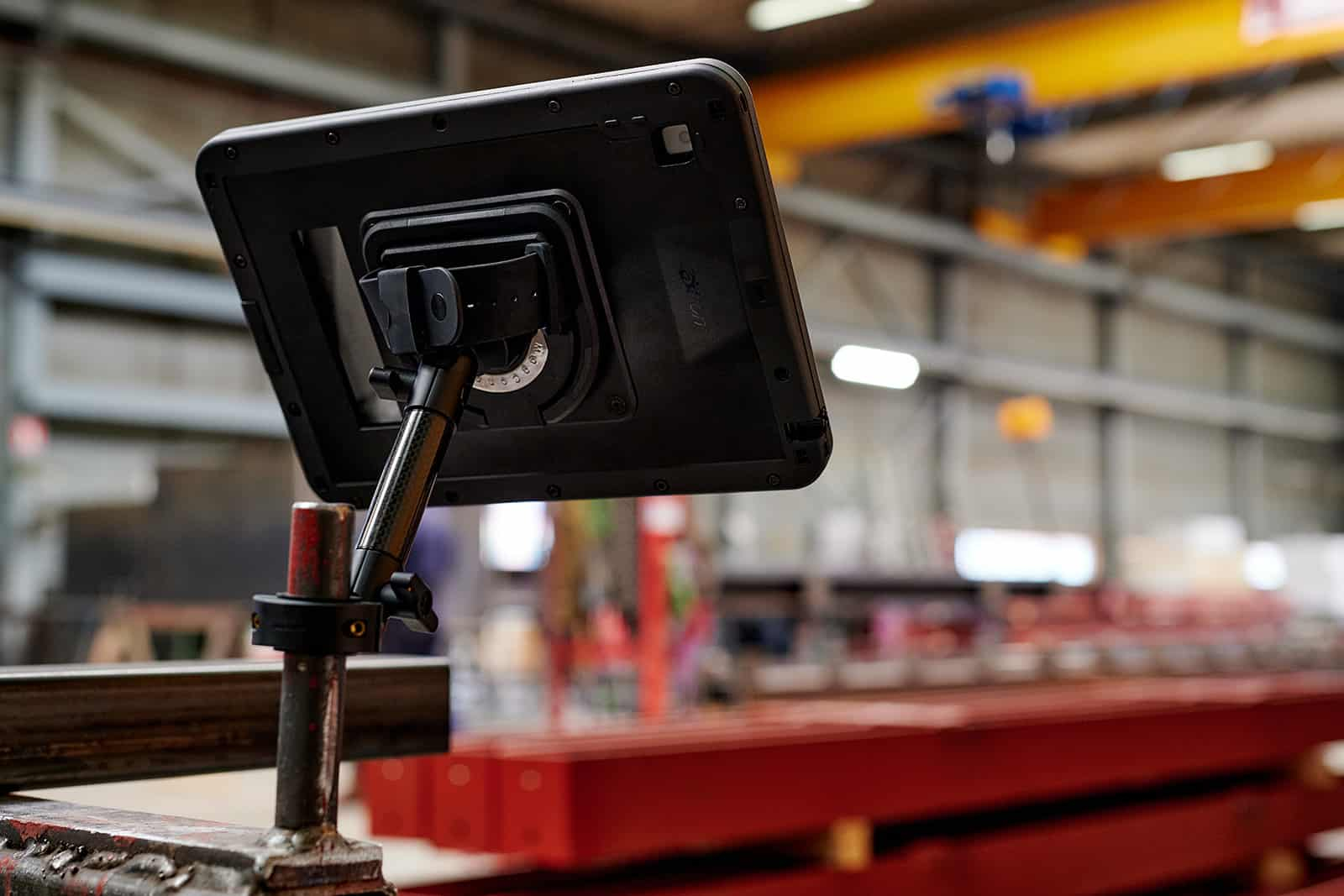 Ein schwarzes Tablet auf einem Ständer in einer Fabrikhalle mit roten Metallträgern im Vordergrund.