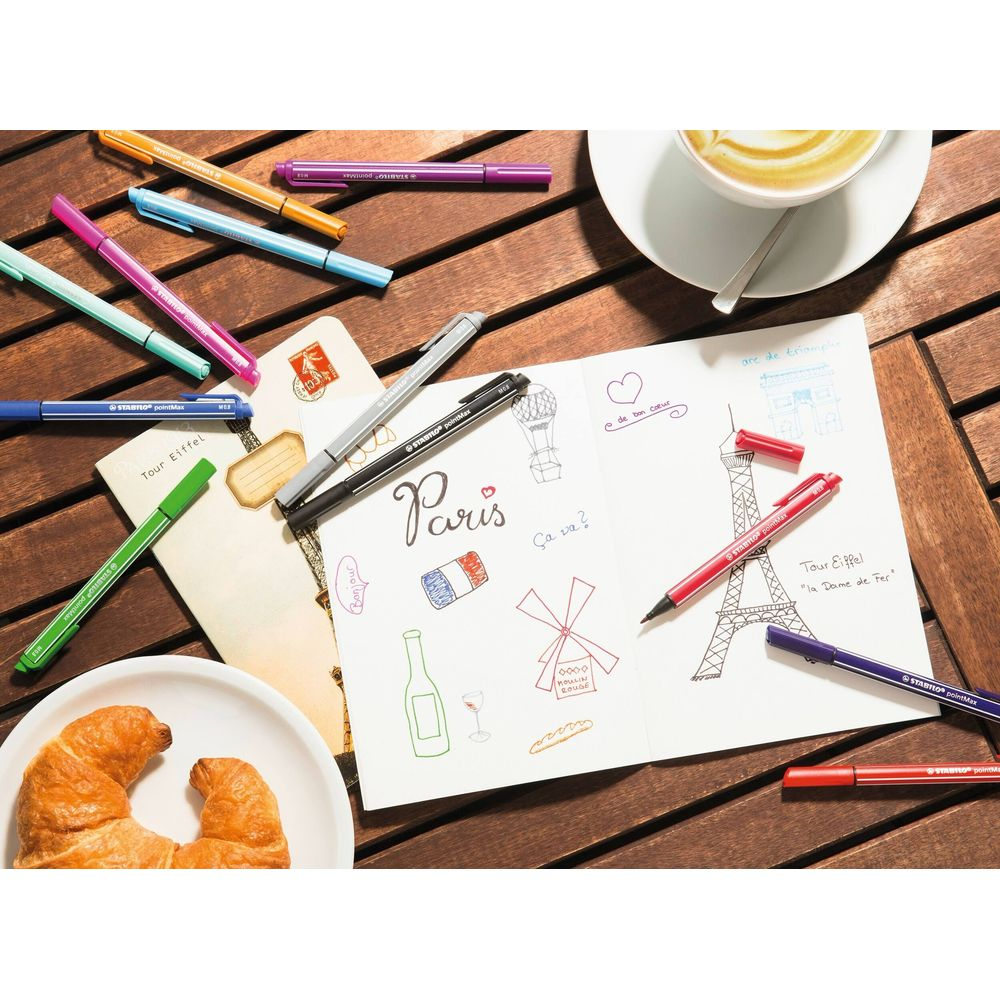 Buntstifte liegen neben einem offenen Notizbuch mit Paris-Zeichnungen und einem Croissant.