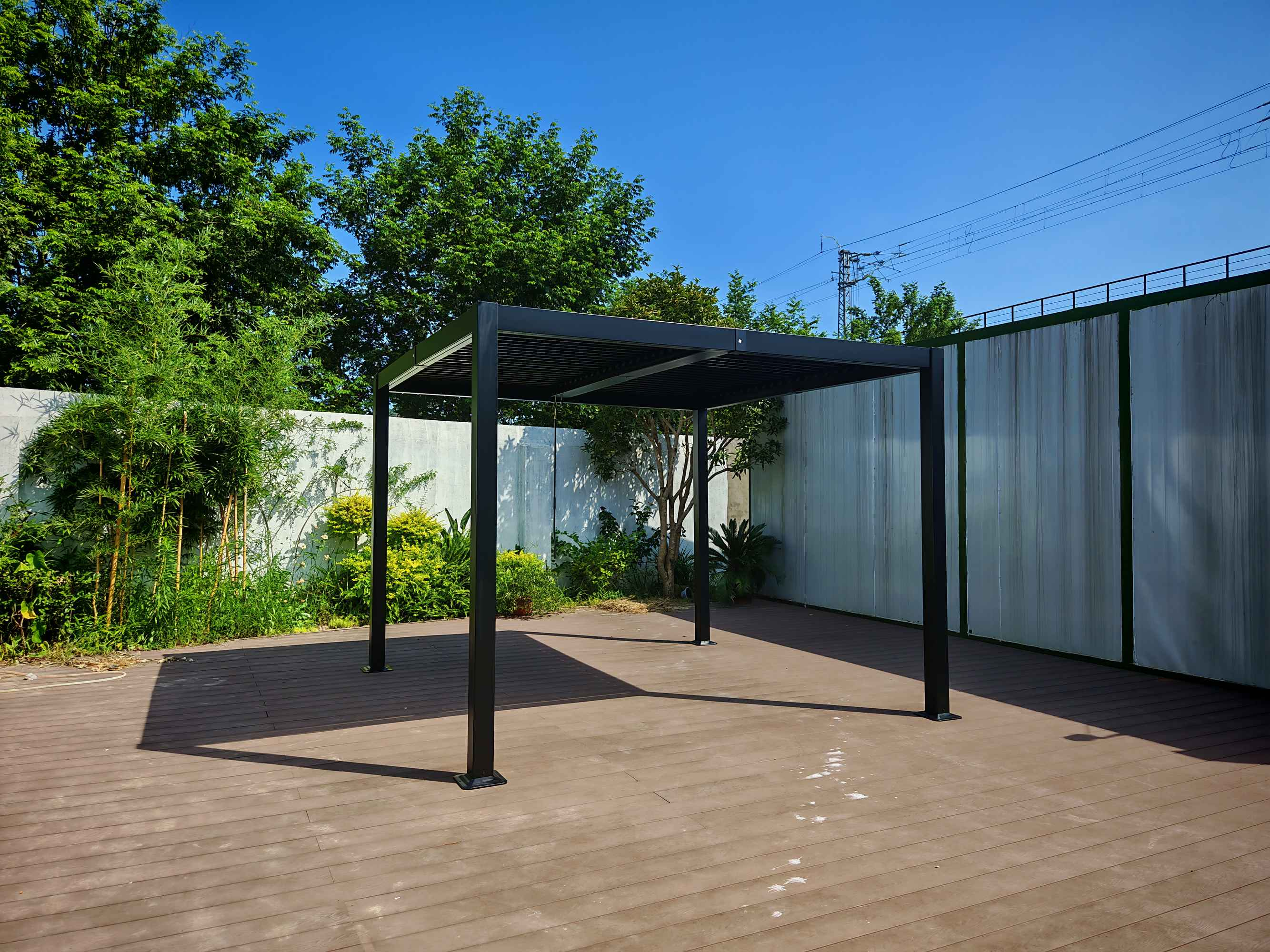 Ein schwarzer Pergola-Pavillon steht auf einer Holzterrasse unter blauem Himmel.