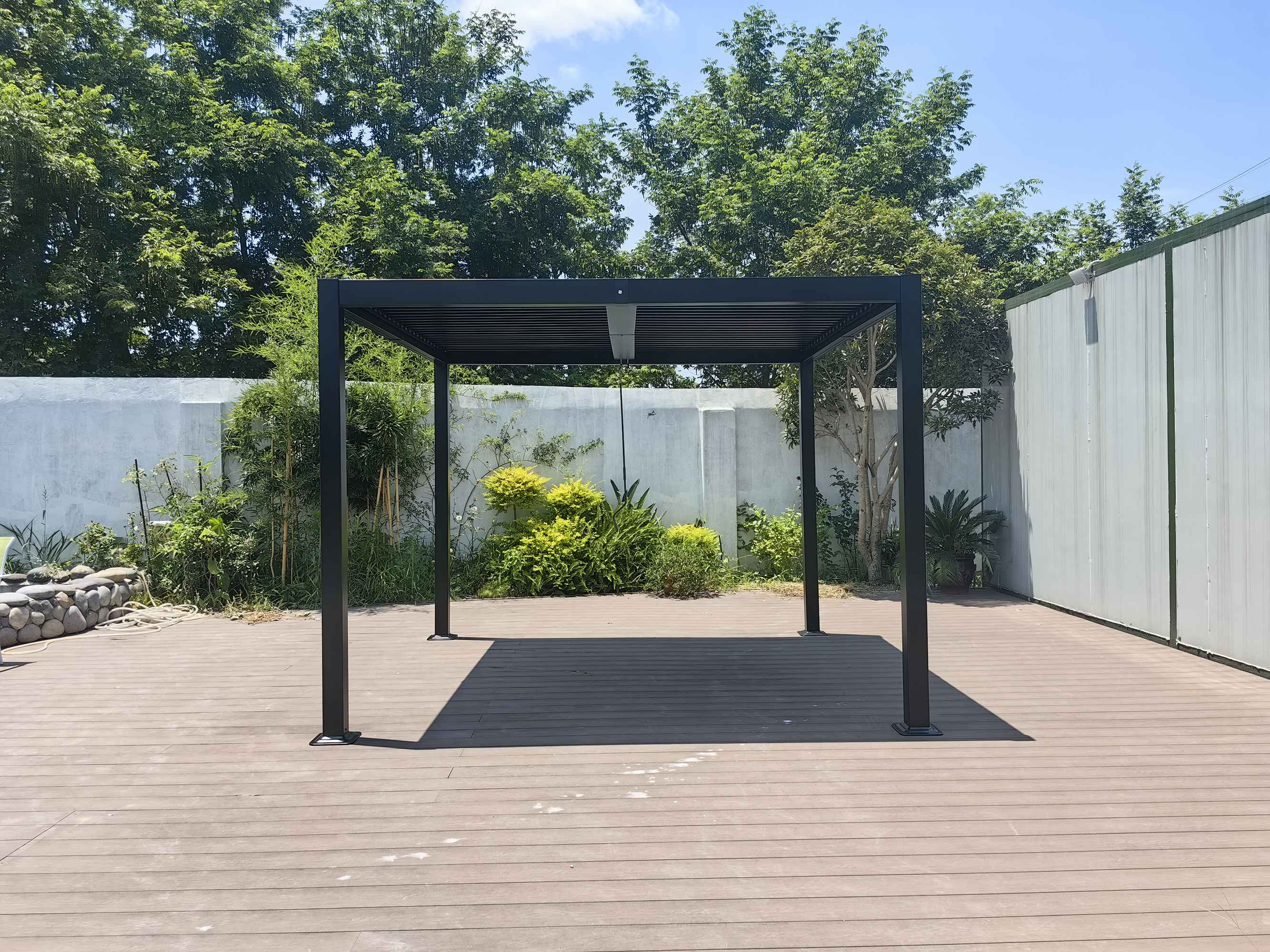 Ein schwarzer Pergola-Pavillon steht auf einer Holzterrasse, umgeben von Grün und einer Mauer.