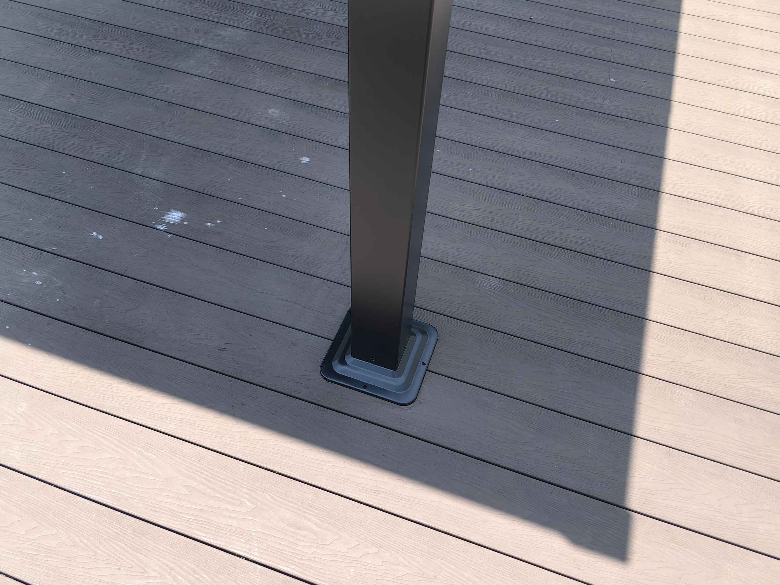 Ein dunkler Pfosten steht auf einem Holzdeck. Schatten fällt von links.