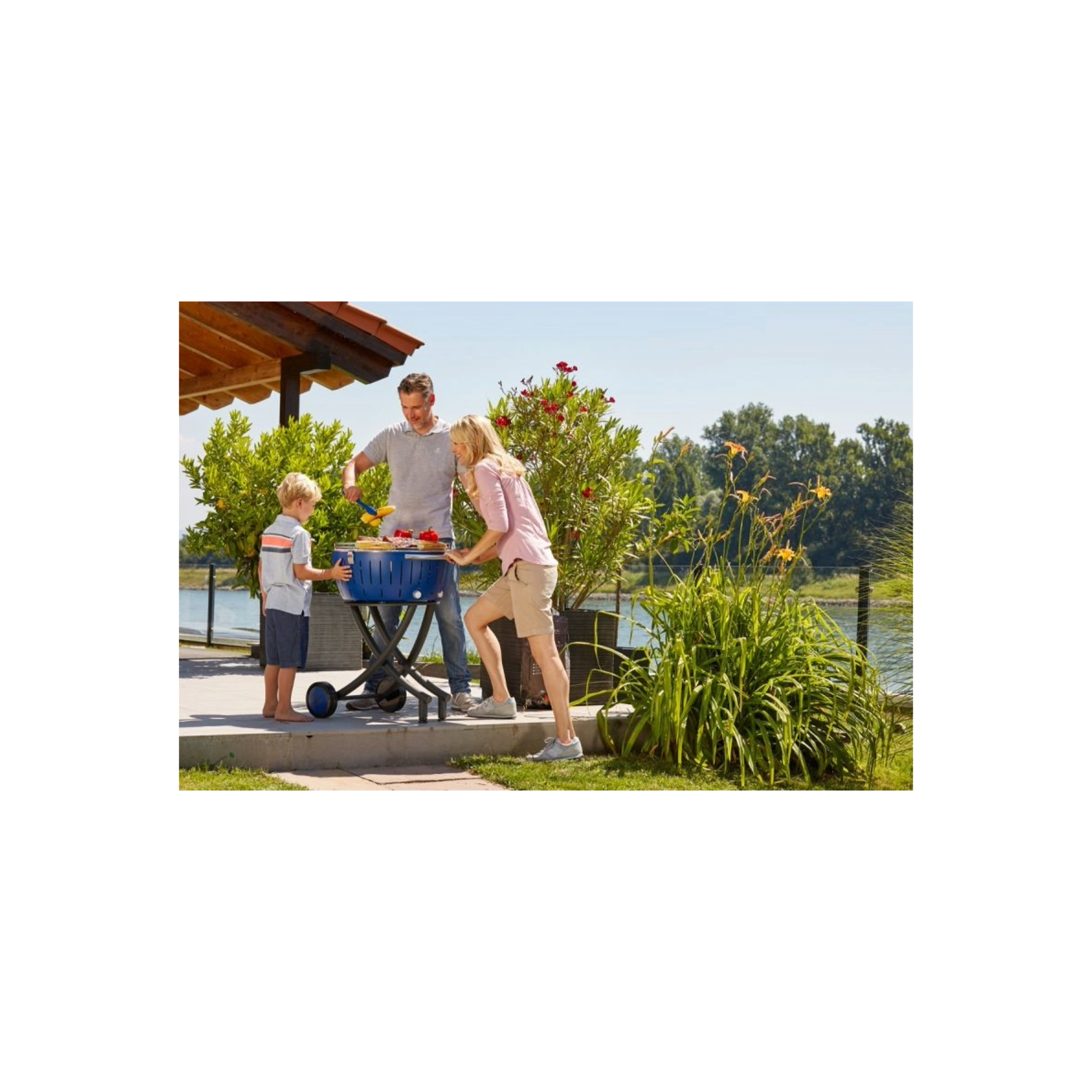 Familie grillt am See unter einem Vordach, Kinder helfen mit.