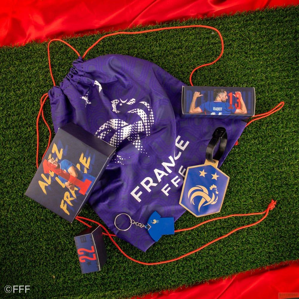 Lila Tasche mit Frankreich-Logo, Fußball-Schlüsselanhänger und Rabiot-Box auf Kunstrasen.