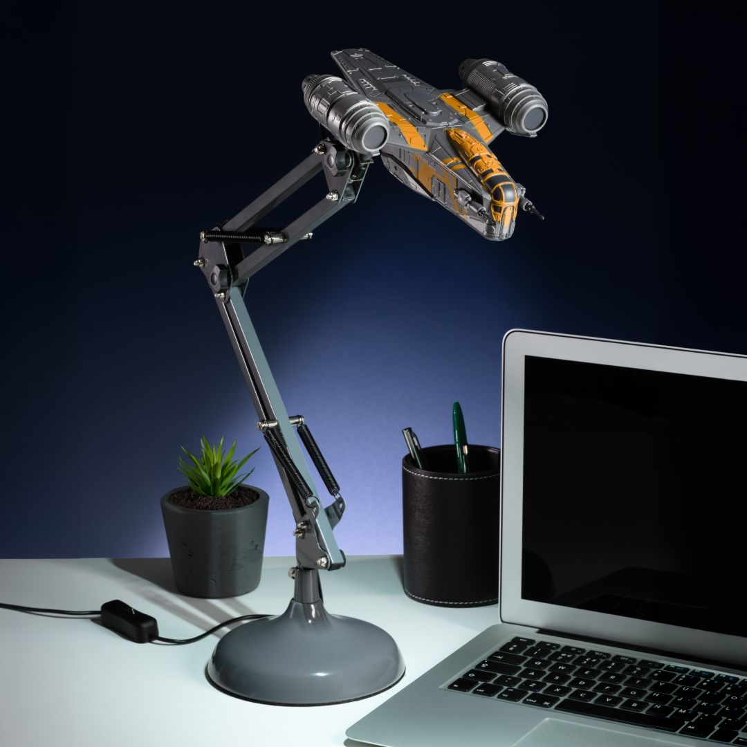 Una lámpara de escritorio gris con forma de nave espacial ilumina un portátil y accesorios de escritorio.