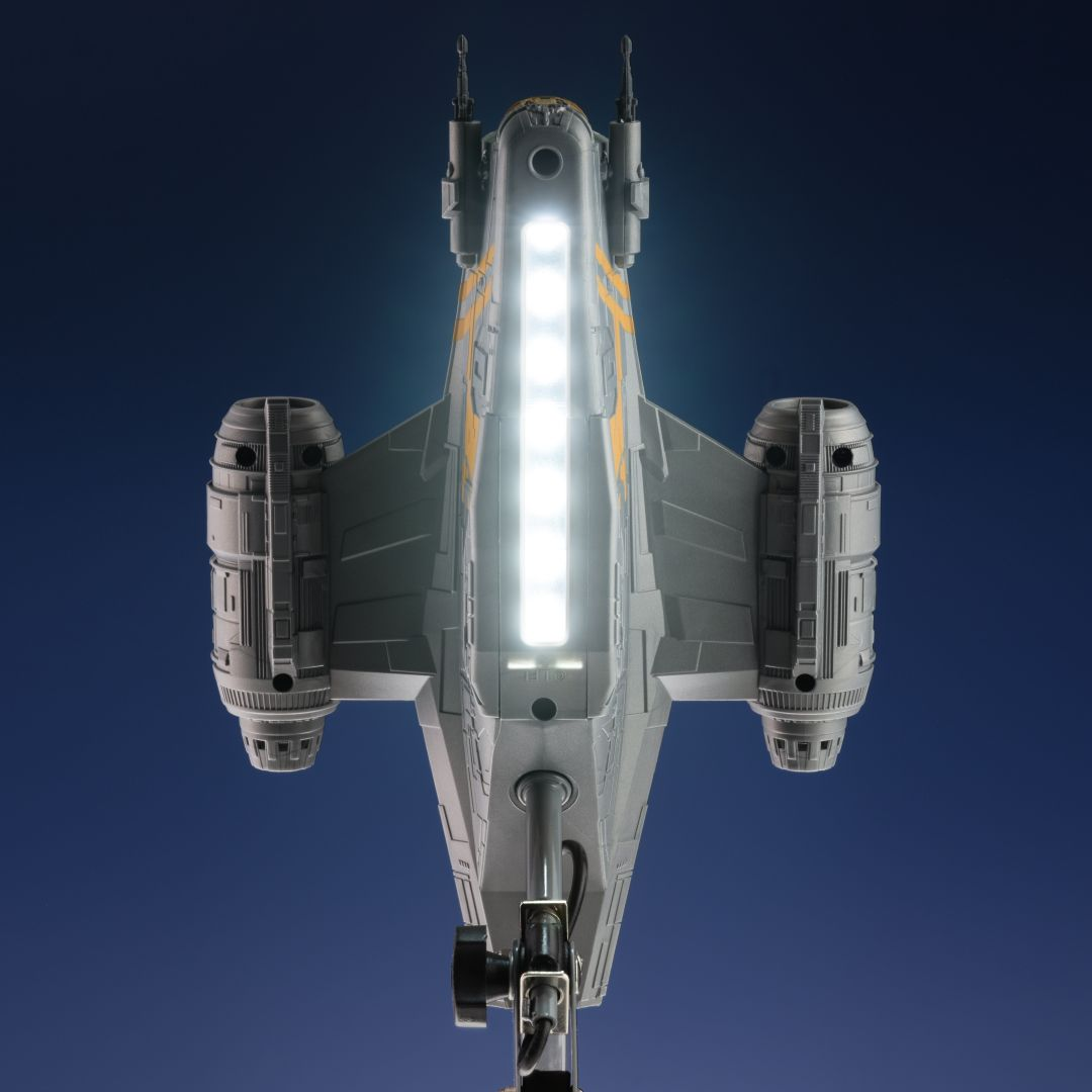 Una nave espacial gris con marcas amarillas y luces brillantes vuela sobre un fondo azul.