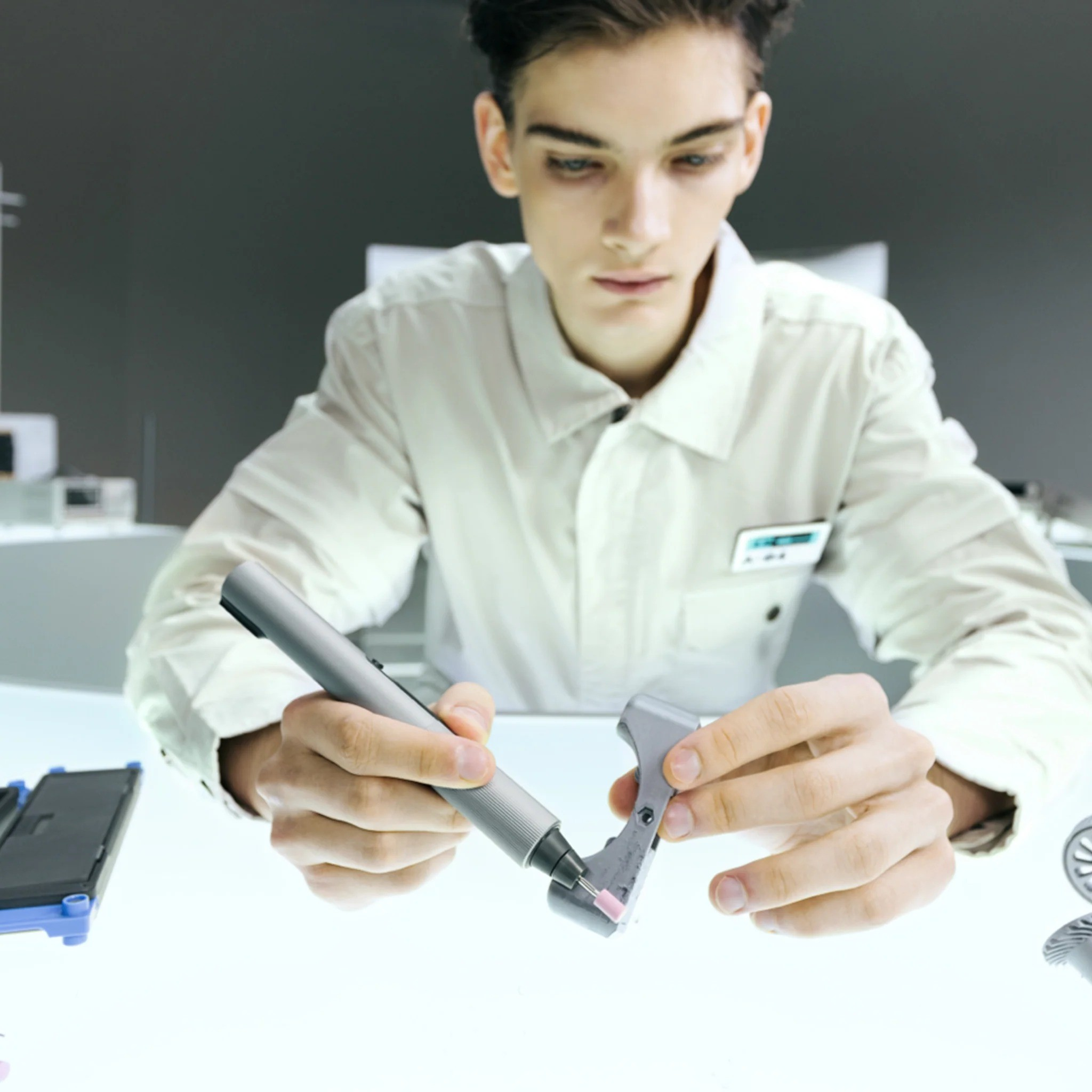 Mężczyzna w białej kurtce laboratoryjnej pracuje nad szarym elementem za pomocą srebrnego narzędzia.