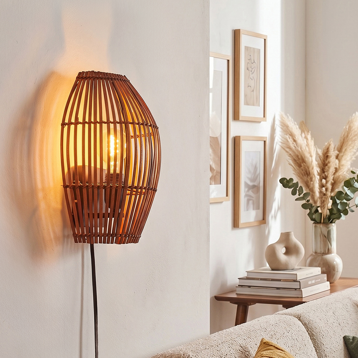 Eine gewebte Bambus-Wandlampe mit Glühbirne beleuchtet eine weiße Wand. Gerahmte Kunst und Pampasgras schmücken den Hintergrund.