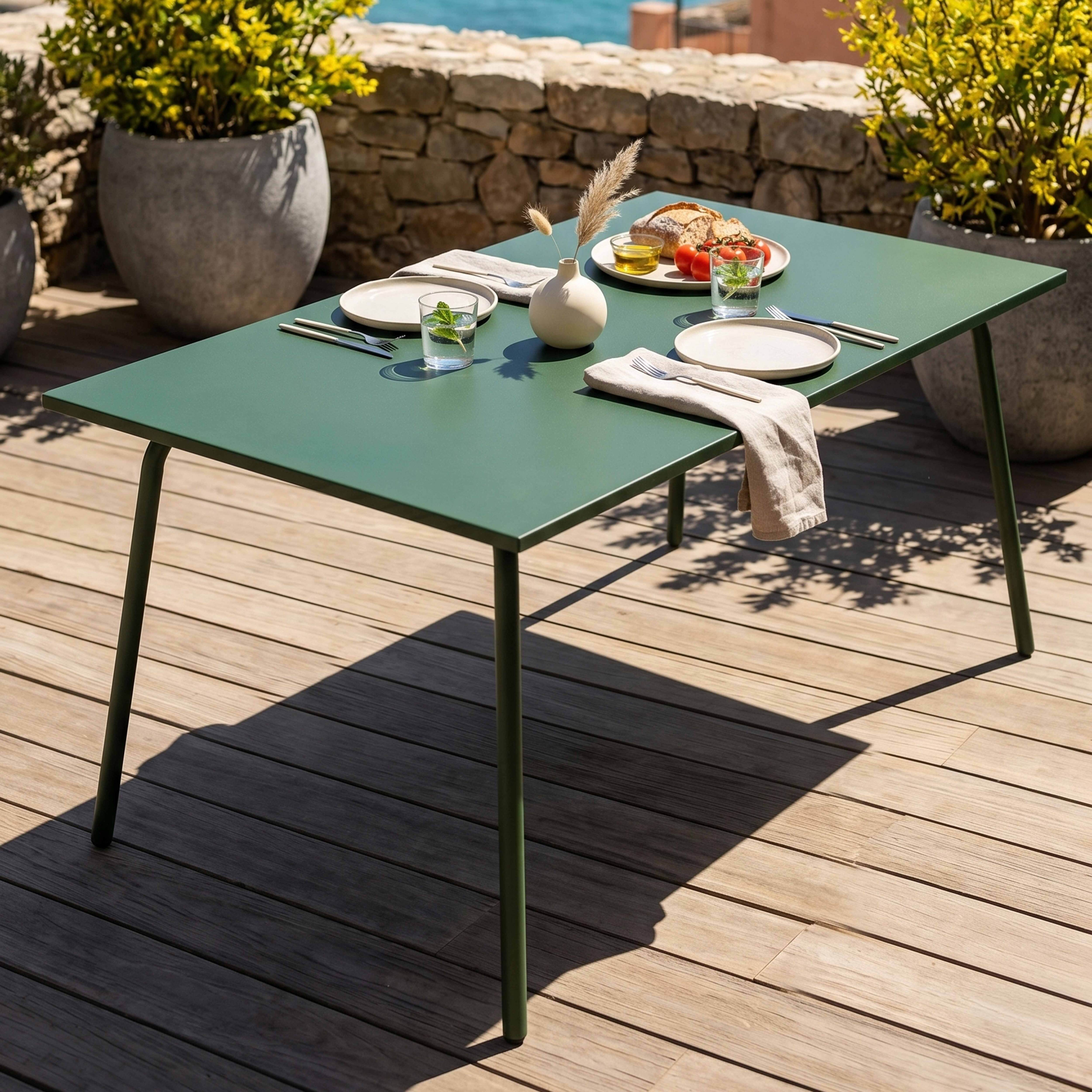 Grüner Tisch mit Essen im Freien, Platten, Brot, Tomaten und Gläsern. Steinmauer und Pflanzen.