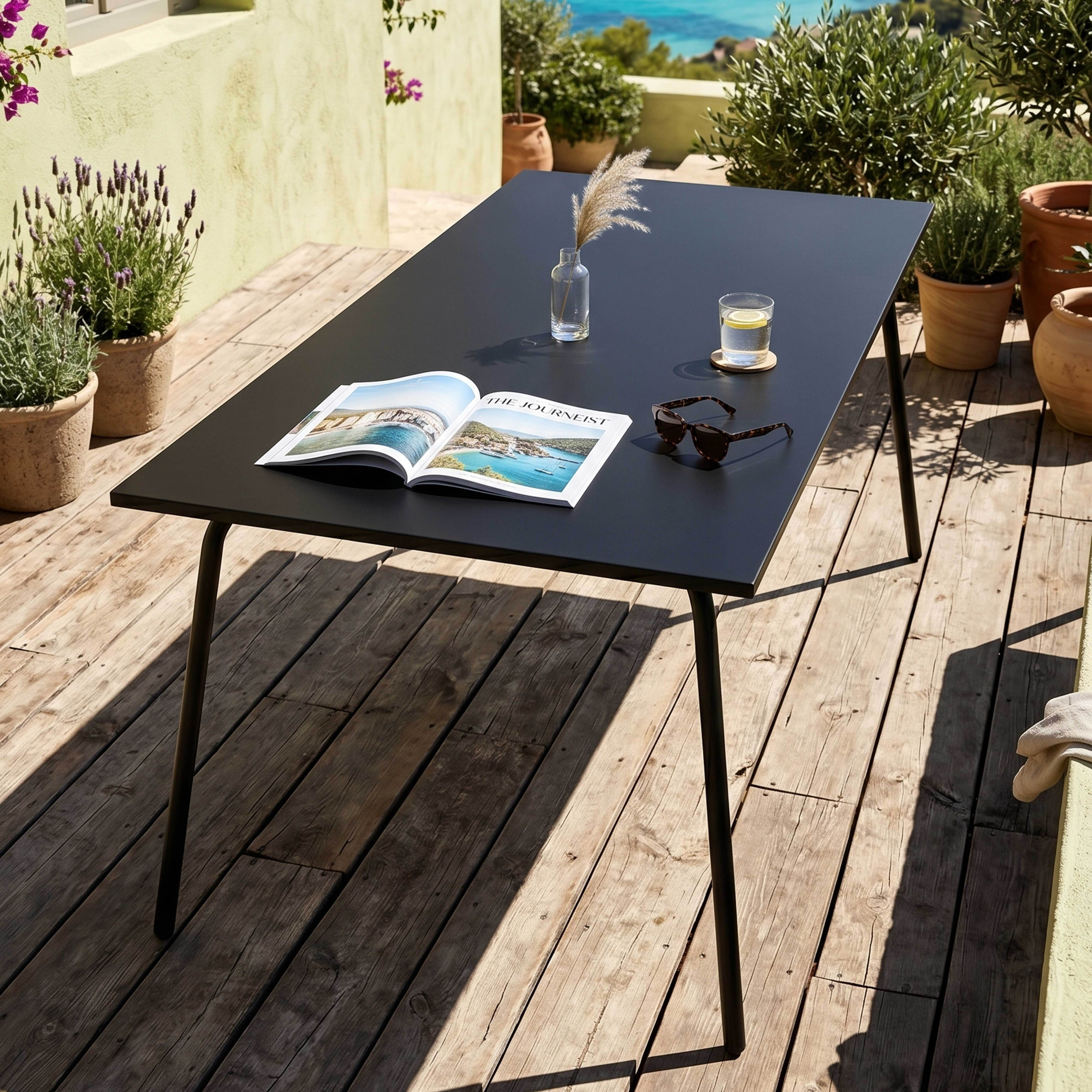 Ein schwarzer Tisch mit einer Zeitschrift, Sonnenbrille und Glas Wasser auf einer Holzterrasse.
