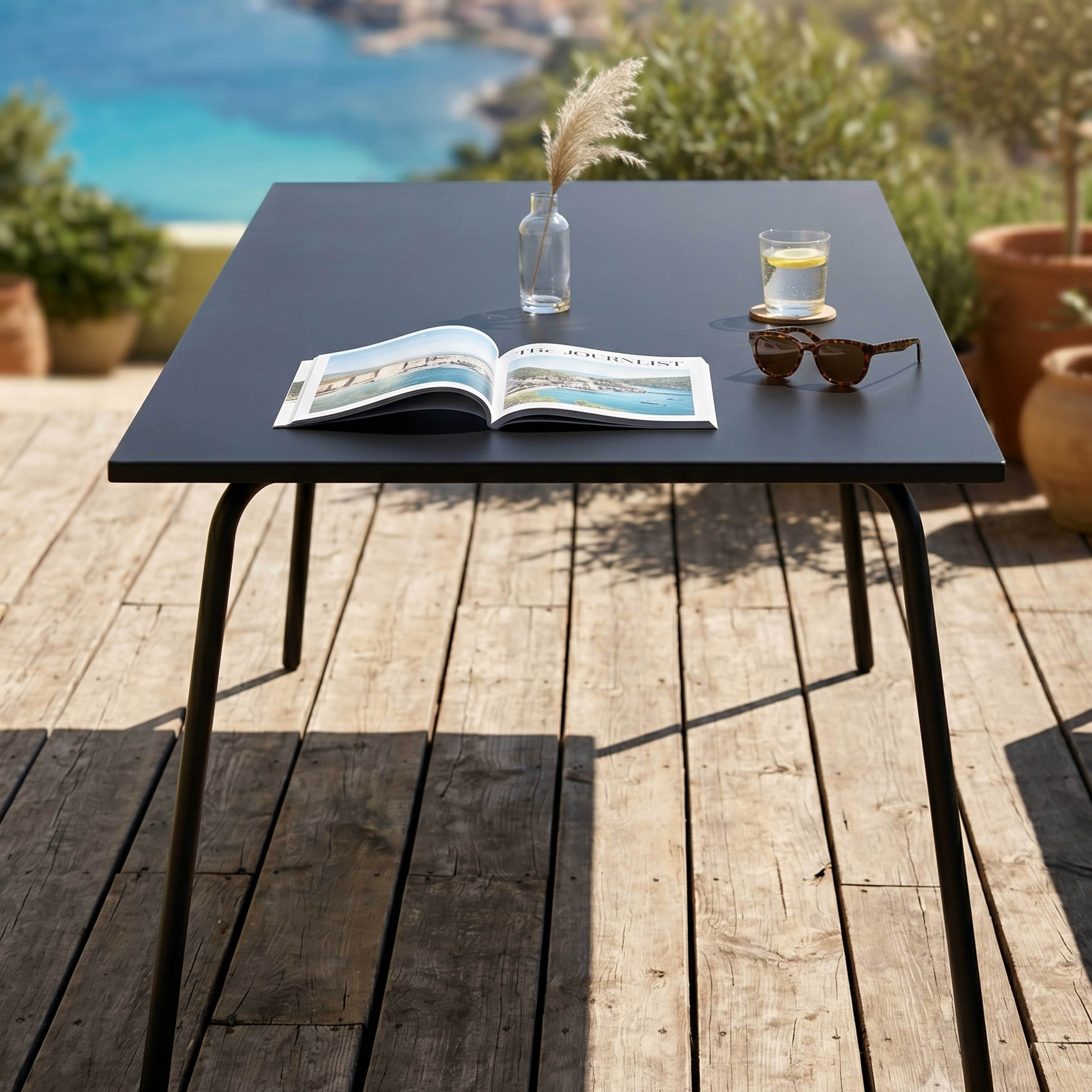 Schwarzer Tisch mit Magazin, Sonnenbrille, Glas und Vase mit Schilf auf Holzdeck.