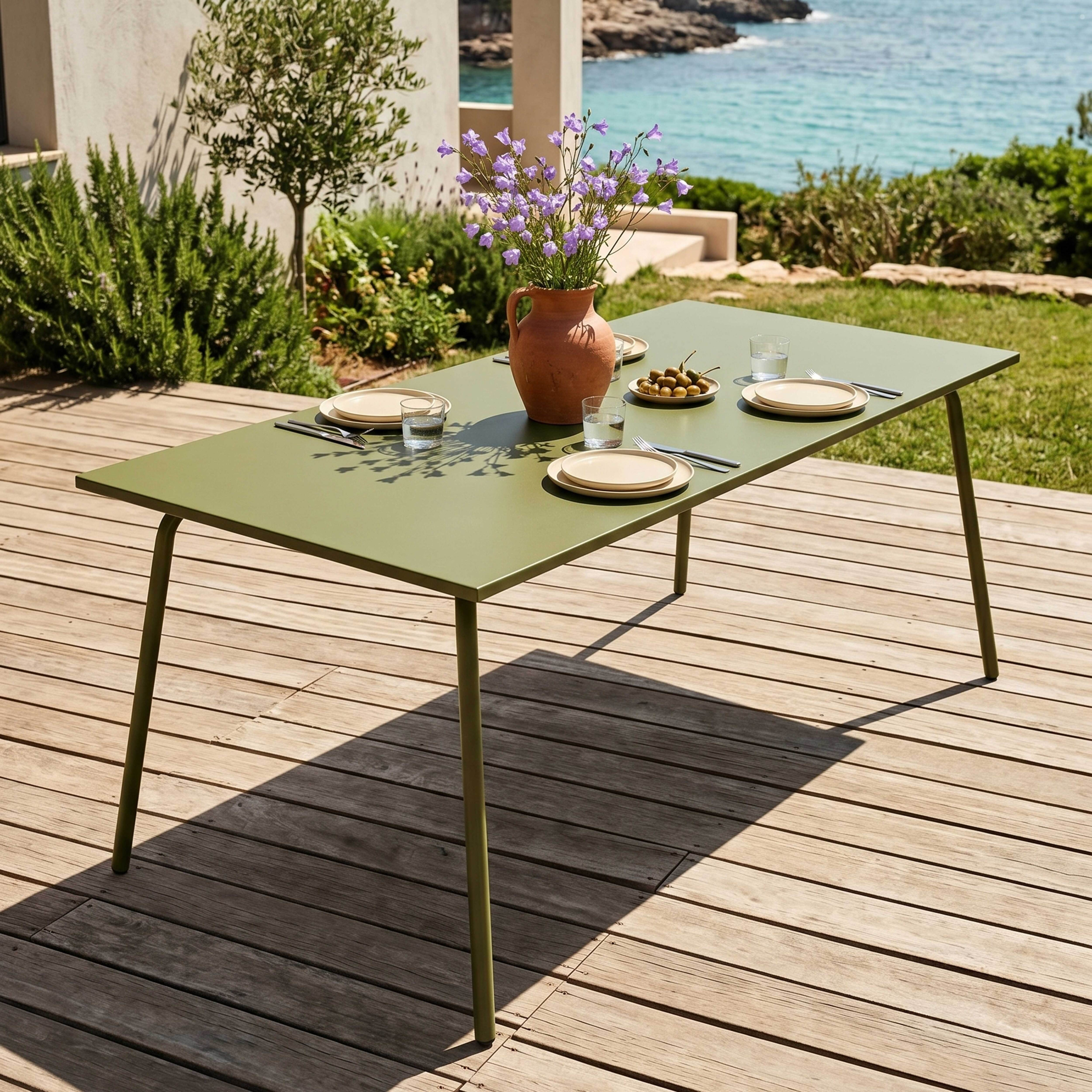 Ein grüner Tisch mit Geschirr und Blumen steht auf einer Holzterrasse am Meer.
