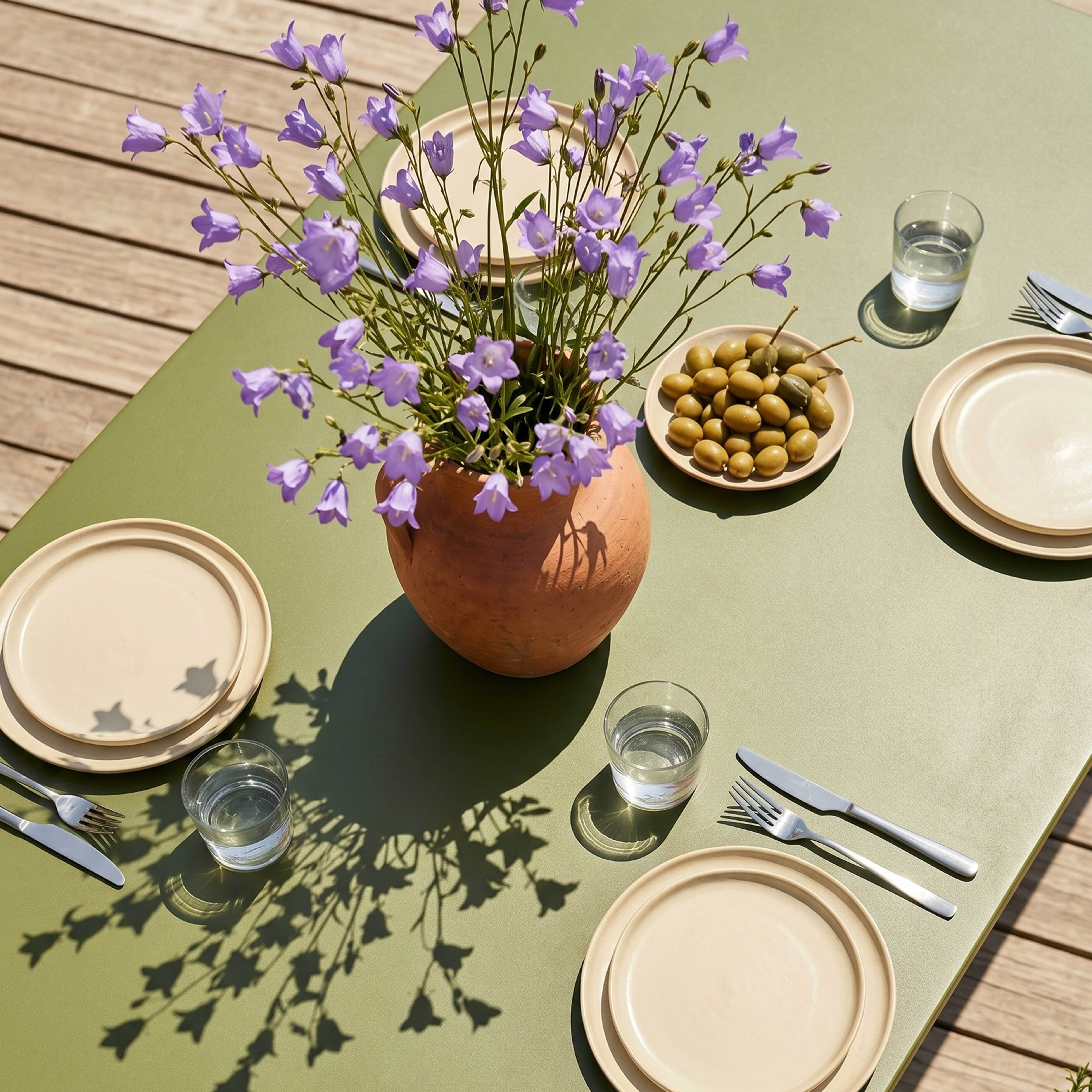 Ein Tisch im Freien mit blau-violetten Blumen in einer Vase, Tellern, Oliven und Gläsern.