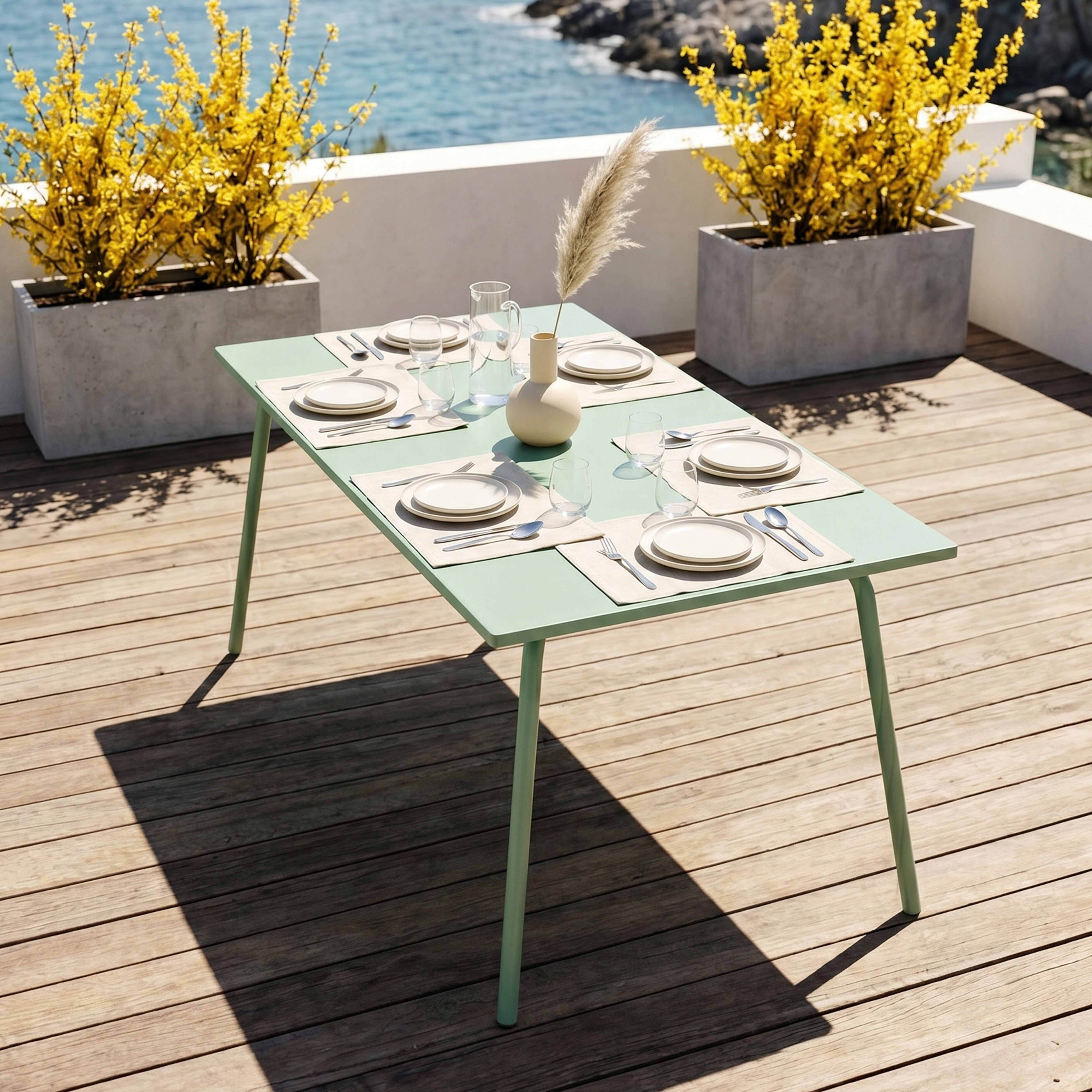Ein grüner Gartentisch mit Gedeck steht auf einer Holzterrasse am Meer.