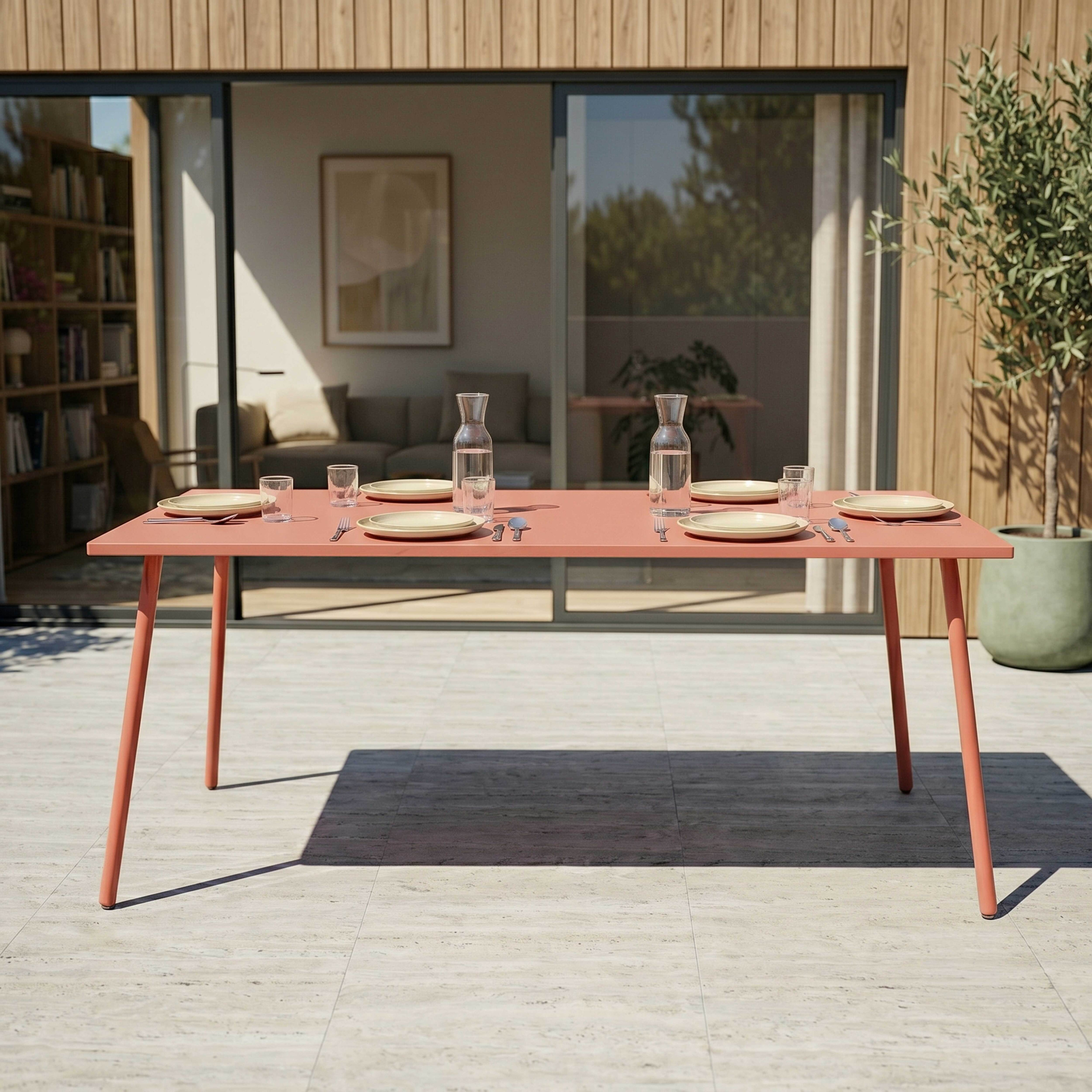 Ein korallenfarbener Tisch mit gedeckter Tafel im Freien, dahinter ein Haus mit Holzverkleidung.