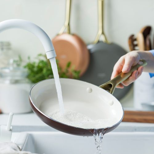 Wasser läuft in eine braune Bratpfanne über einem Spülbecken, mit Töpfen im Hintergrund.