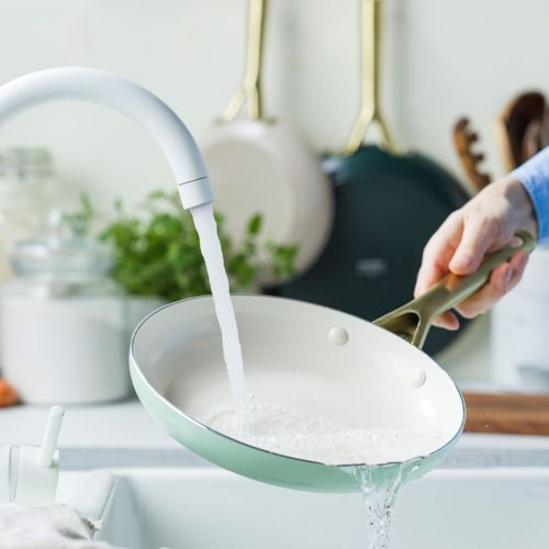 Eine hellgrüne Bratpfanne wird in einer Küchenspüle unter einem weißen Wasserhahn gespült.