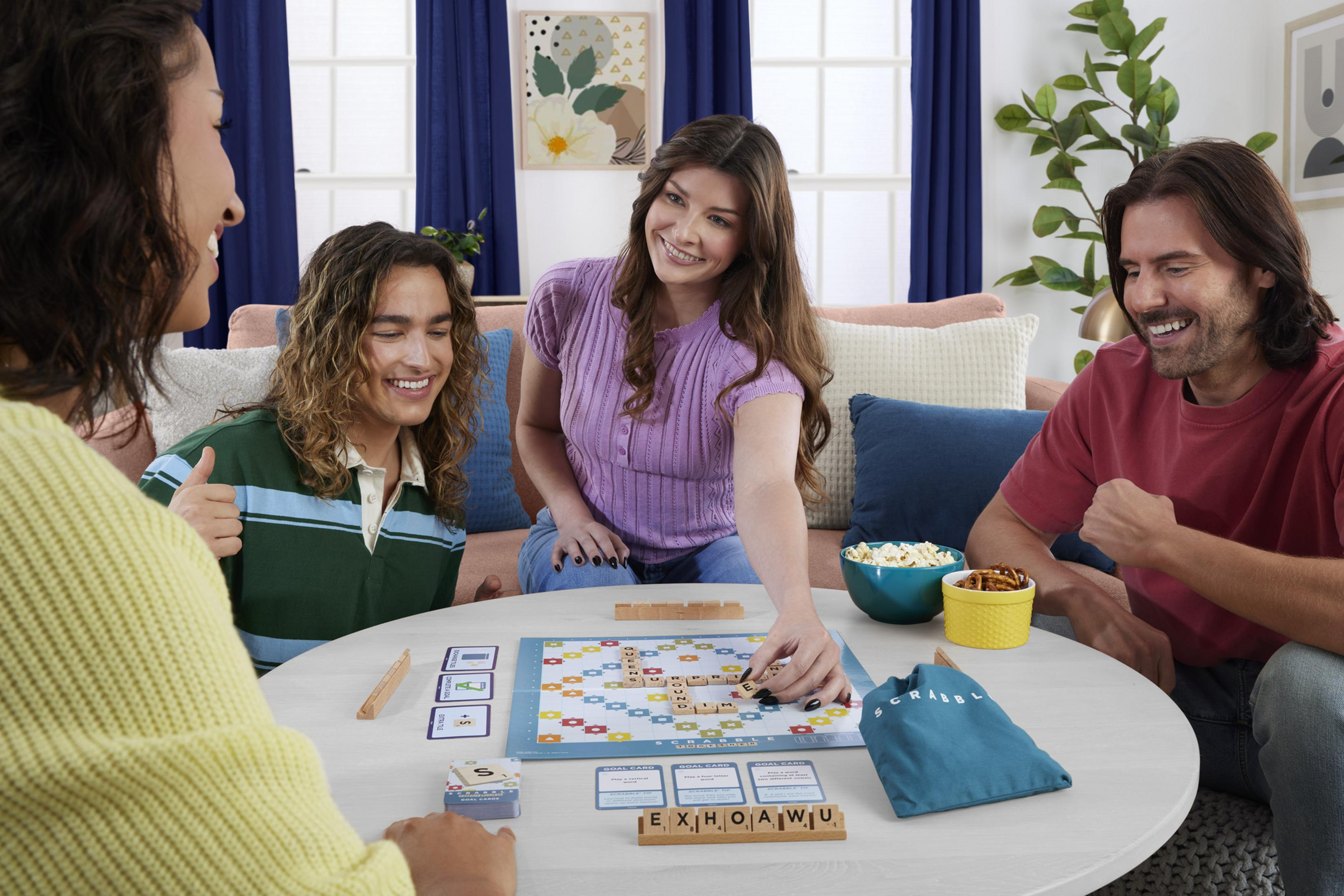 Vier Personen spielen Scrabble an einem runden Tisch mit Snacks. Sie lachen und tauschen Spielsteine.