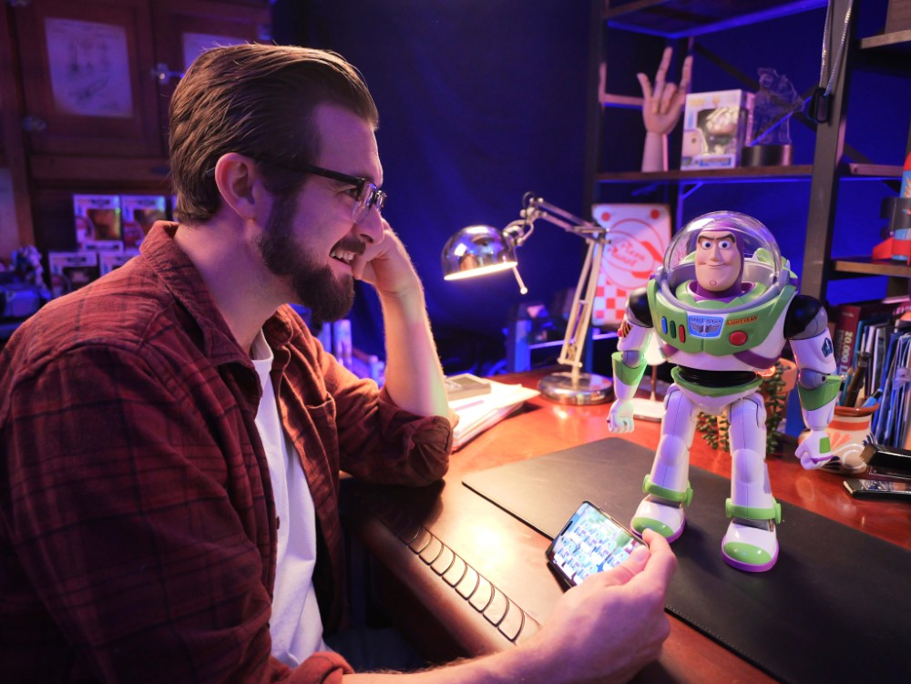 Hombre con camisa roja sonríe, mira su teléfono, juguete Buzz Lightyear en escritorio.