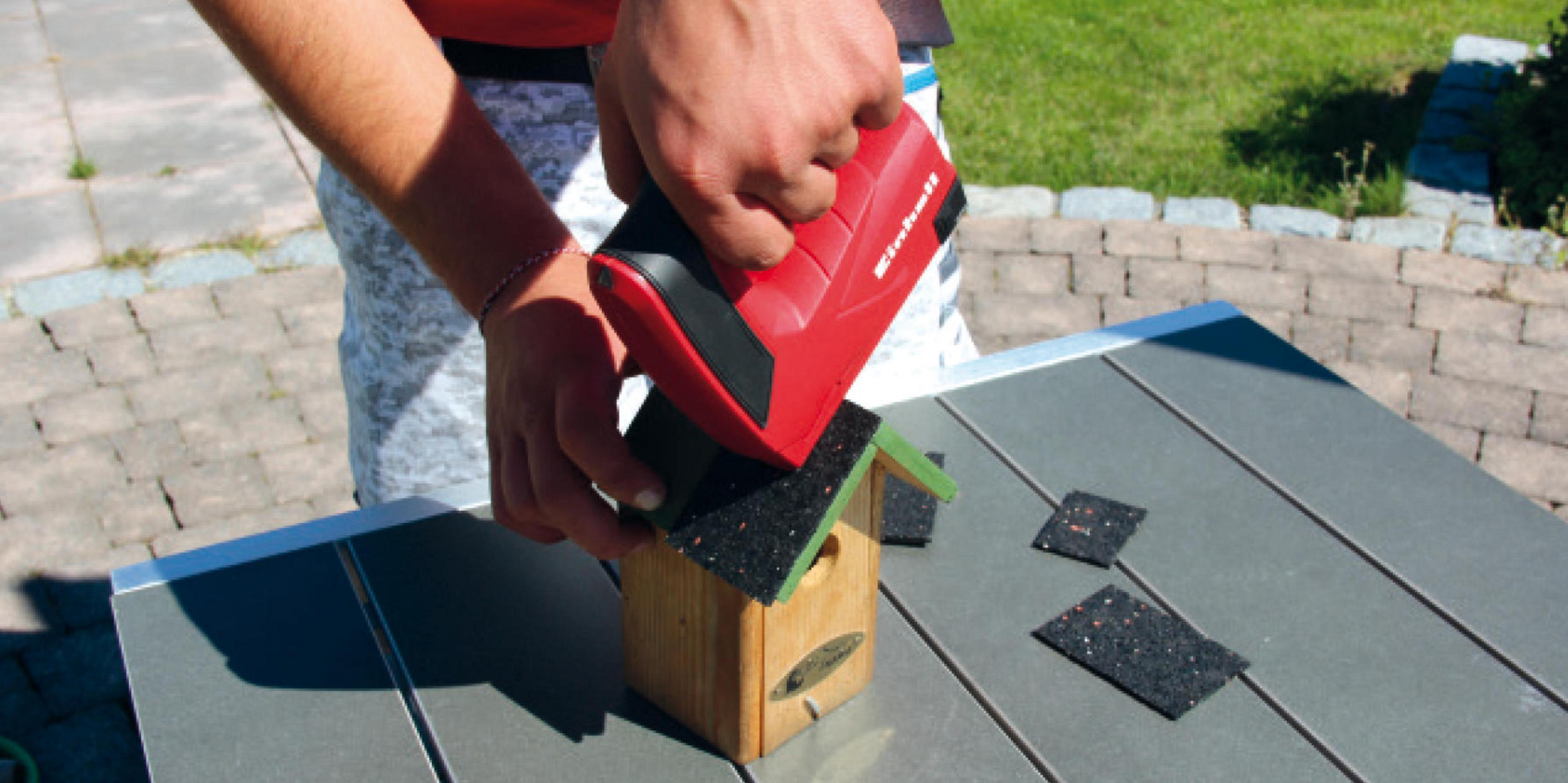 Una persona attacca un tetto a una casetta di legno per uccelli con un dispositivo rosso.