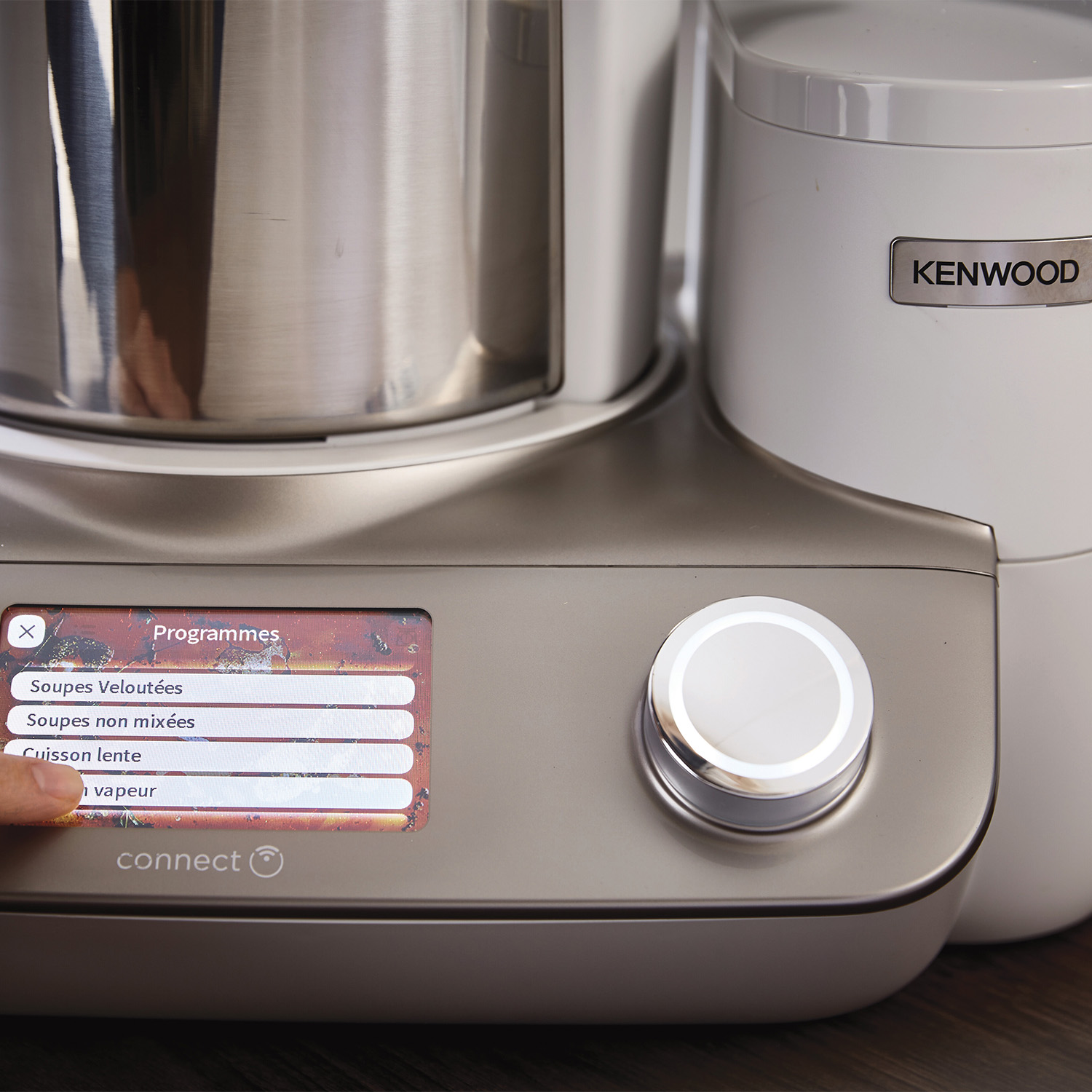 Kenwood Kochmaschine Bildschirm zeigt Programme: Suppen, Schongaren, Dampf. Ein Finger berührt den Bildschirm.