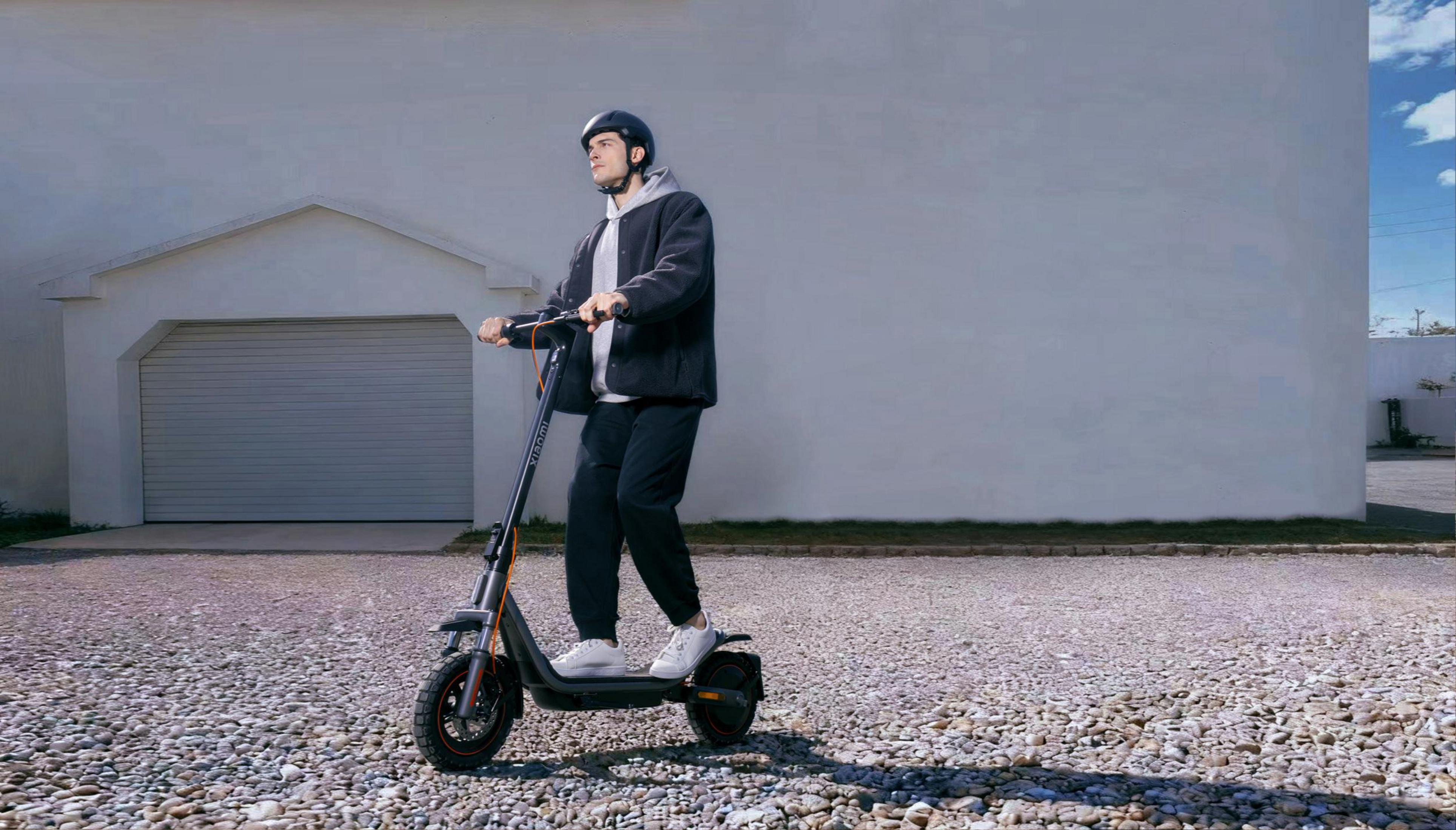 Mann mit Helm fährt E-Scooter vor Garage, Kiesweg, blauer Himmel