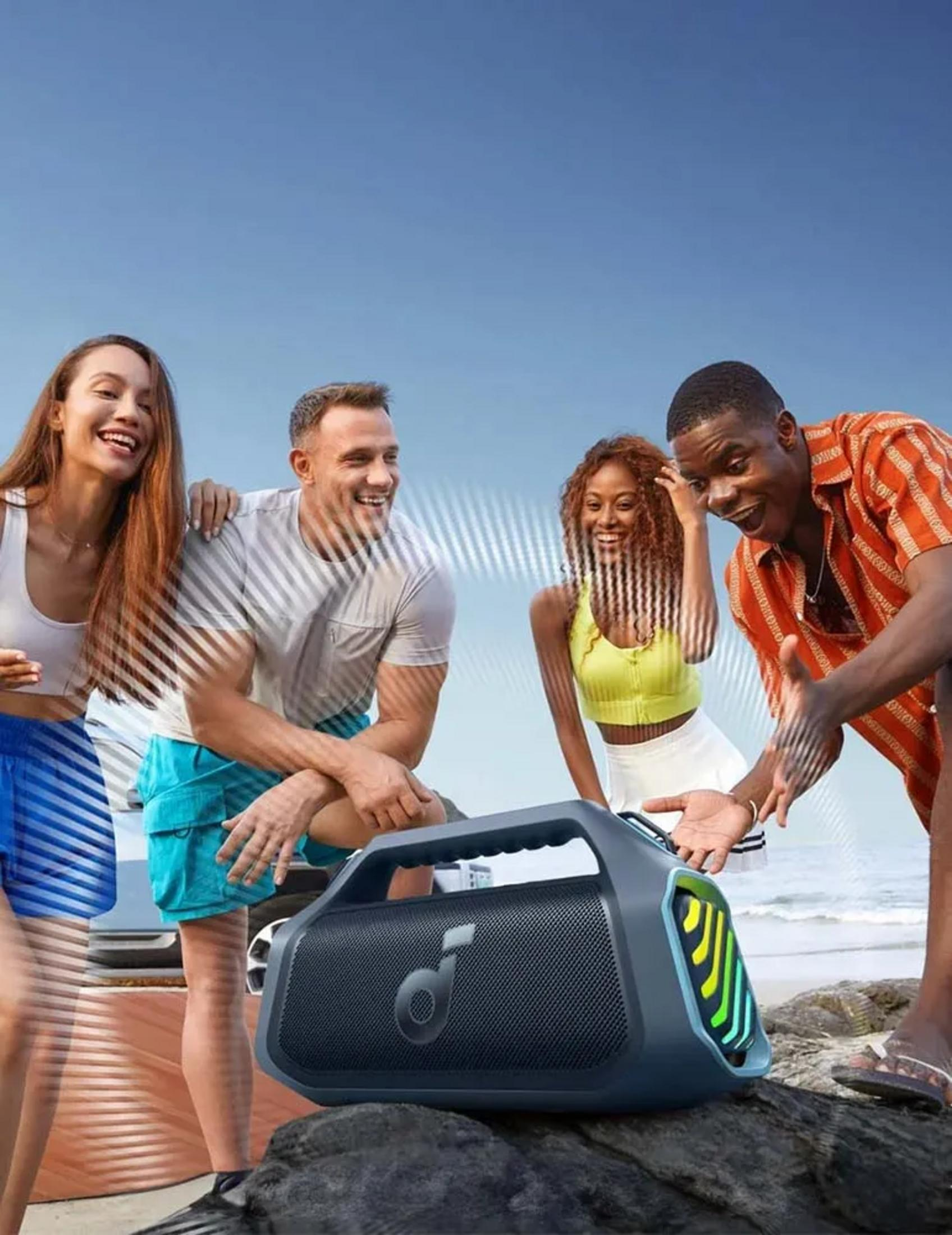 Quattro amici ridono su una spiaggia con uno speaker portatile. Cielo blu e oceano.