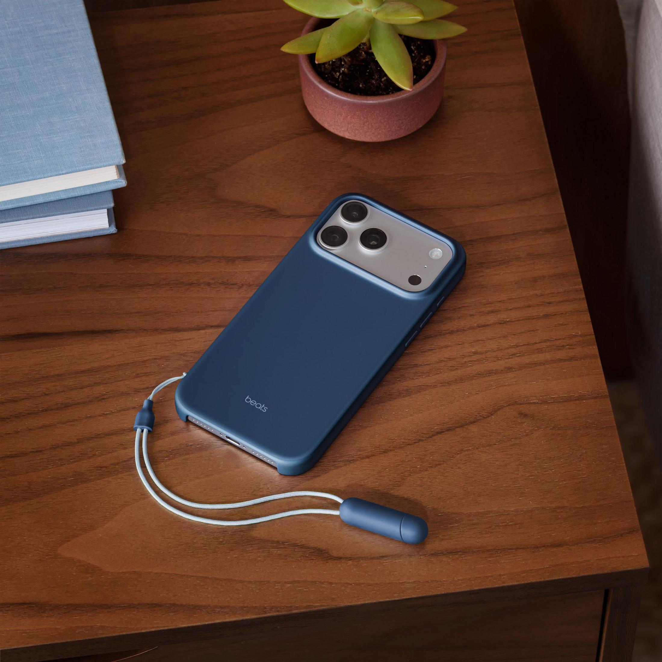 Ein blaues Handy mit Lanyard liegt auf einem Holztisch neben Büchern und einer Pflanze.