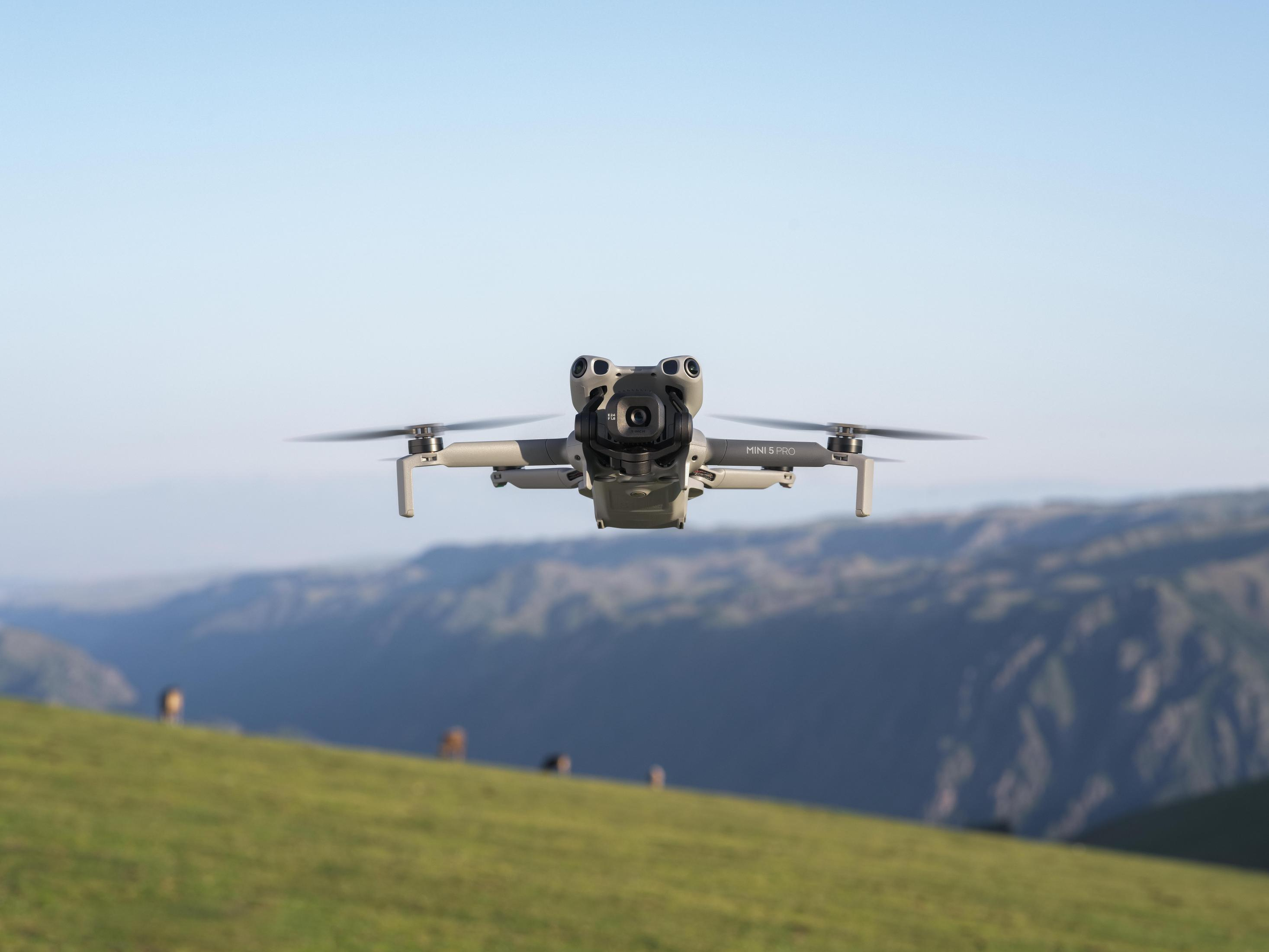 Un dron gris vuela sobre un paisaje de colinas verdes con montañas al fondo.