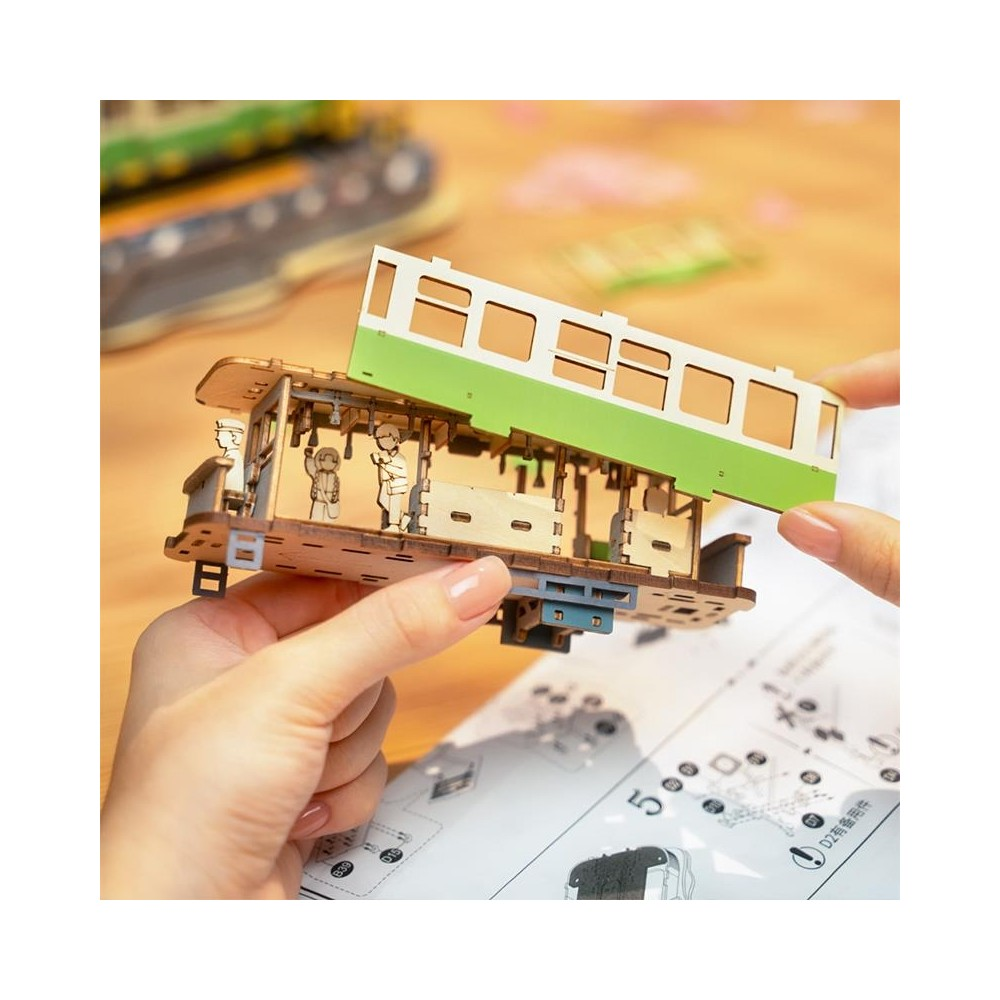 Hände montieren ein Holzmodell einer grünen und weißen Straßenbahn mit kleinen Figuren.