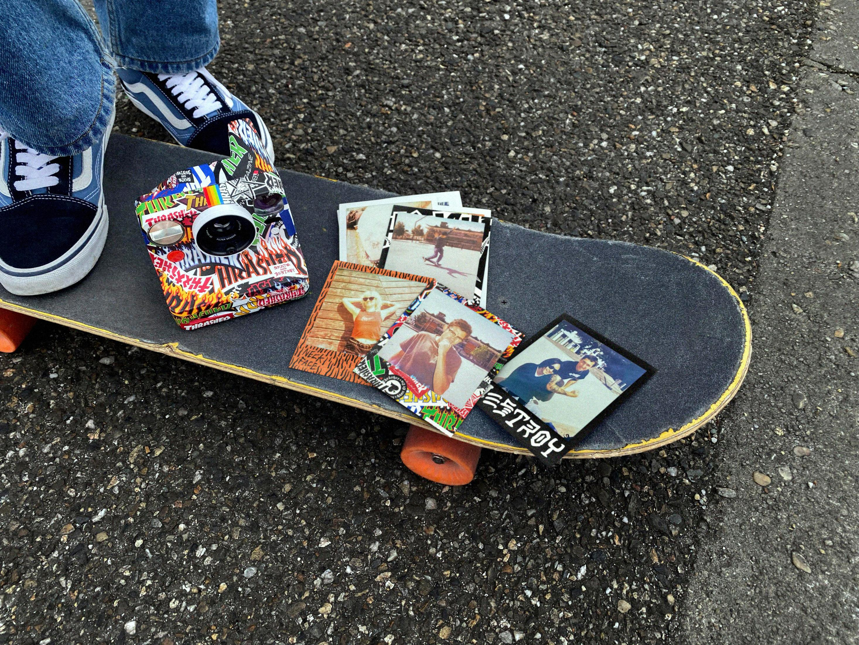 Uno skateboard con adesivi, foto e una fotocamera sopra, con jeans blu e scarpe Vans.