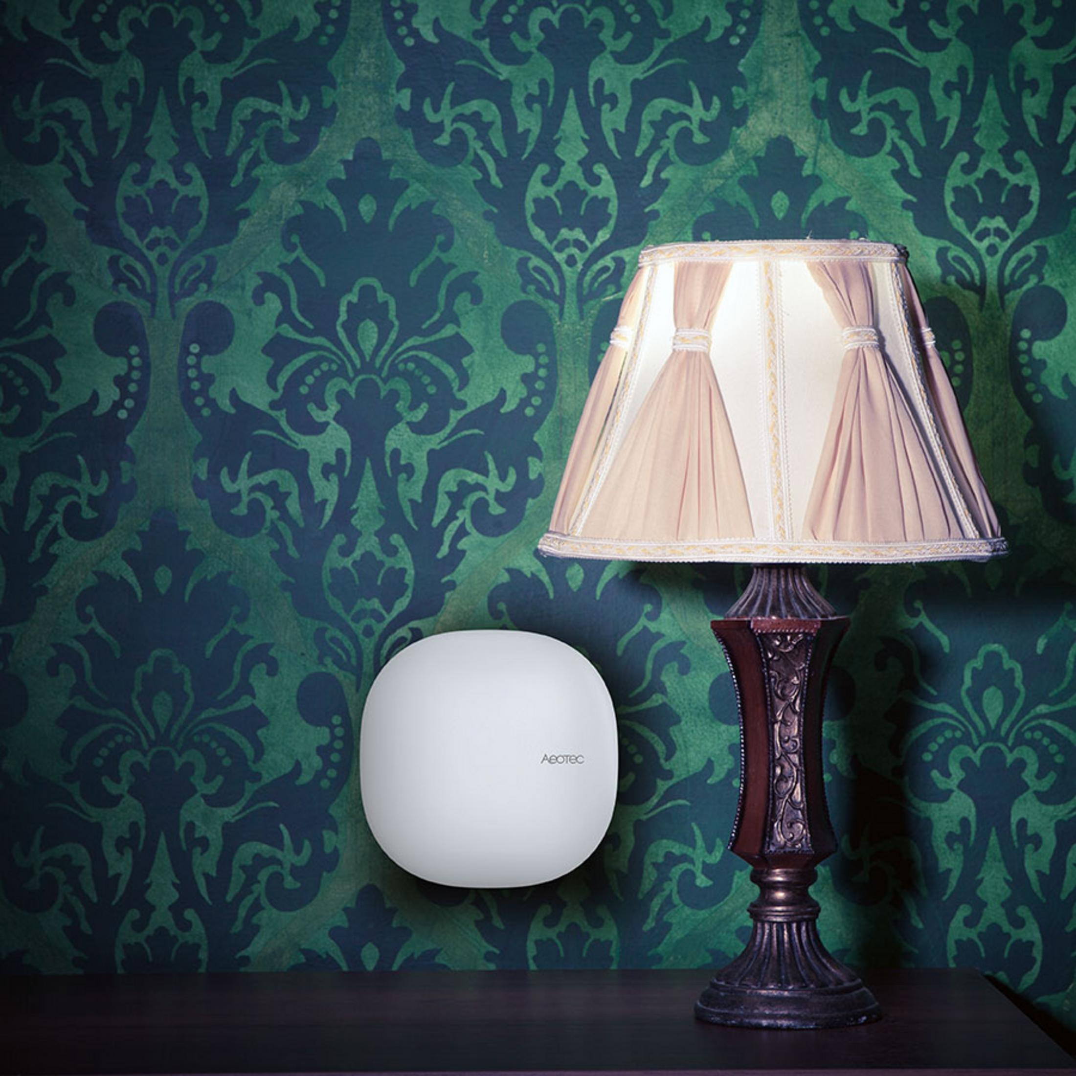 Ein weißes, ovales Smart-Home-Gerät ist neben einer Tischlampe an einer grünen Damasttapete montiert.
