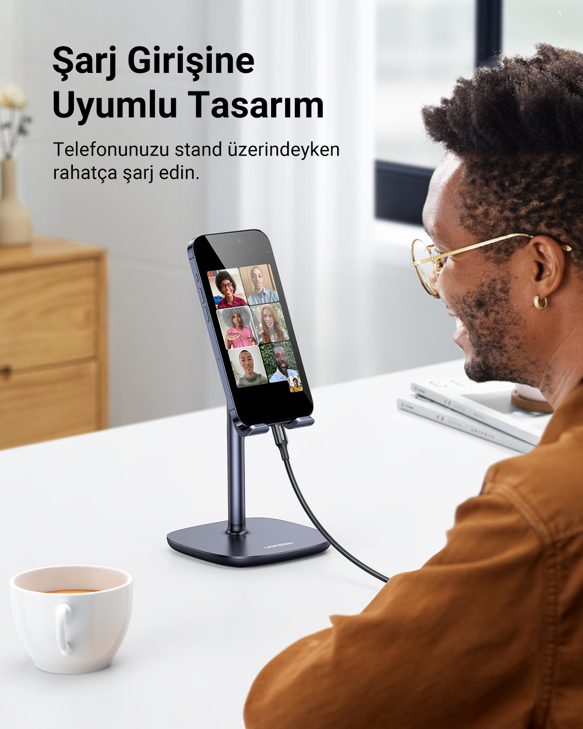 Gözlüklü adam telefonu stantta tutuyor, şarj ediyor. Yanında kahve fincanı var.