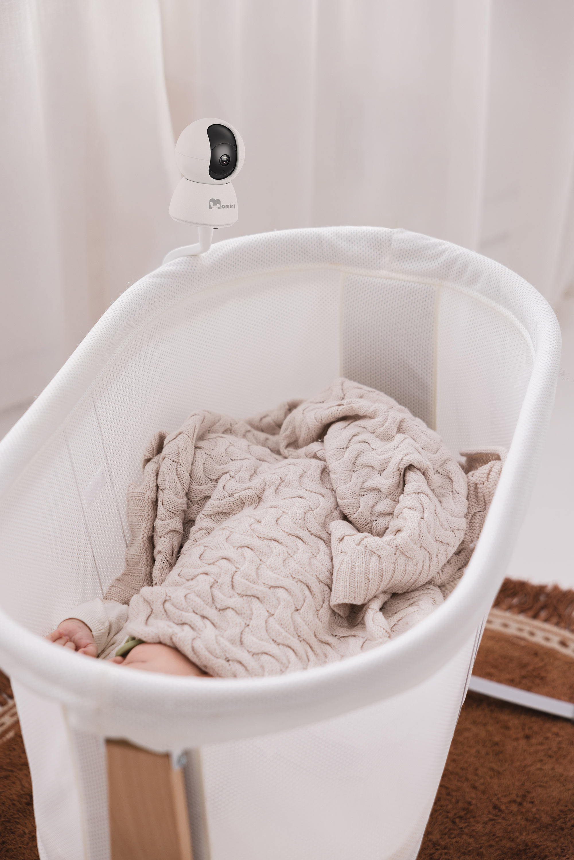 Un bambino giace in una culla bianca, coperto da una coperta lavorata a maglia beige. Una telecamera è fissata sopra.
