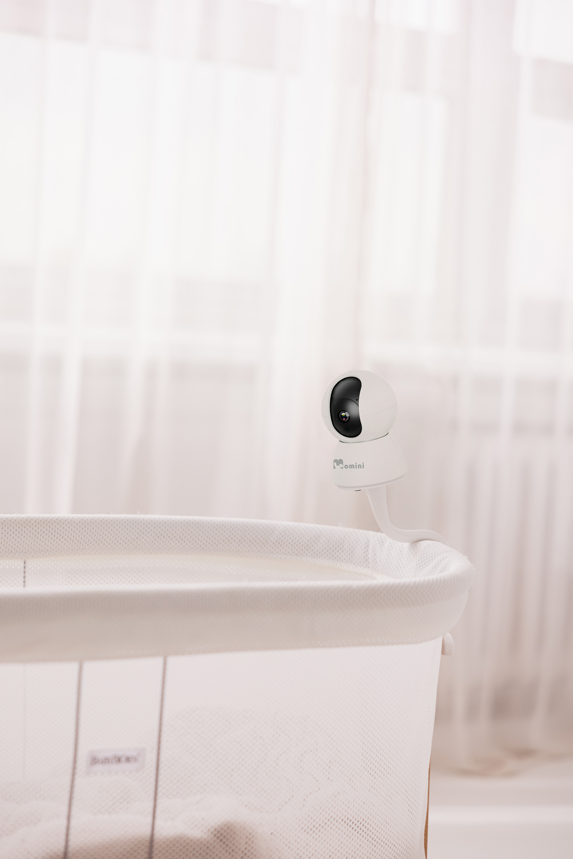 Il braccio di una telecamera baby monitor bianca si estende sopra una culla bianca con una finestra sullo sfondo.