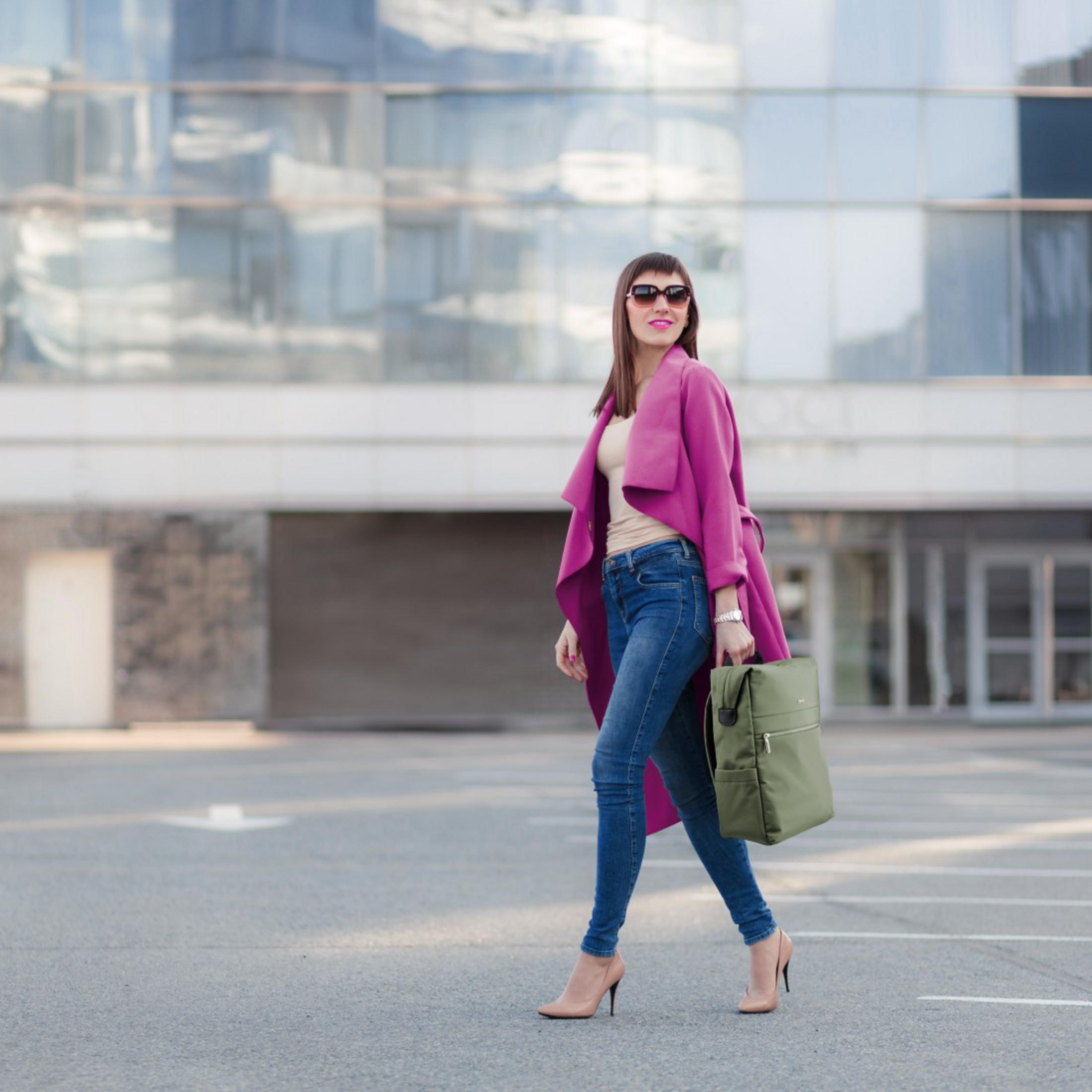 Donna in jeans, cappotto rosa, occhiali da sole e borsa cammina davanti edificio moderno.