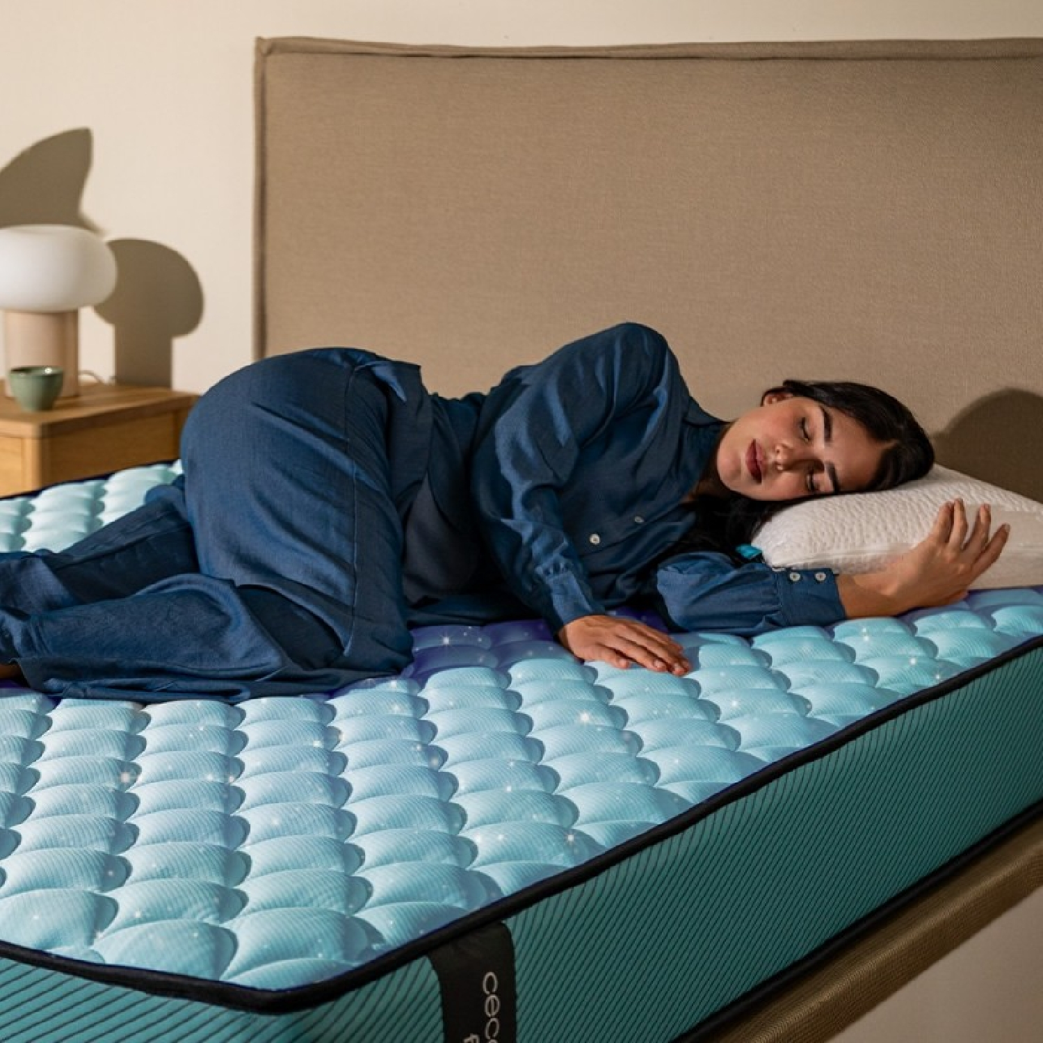 Una mujer de mezclilla duerme en un colchón azul junto a una mesita de noche con lámpara.