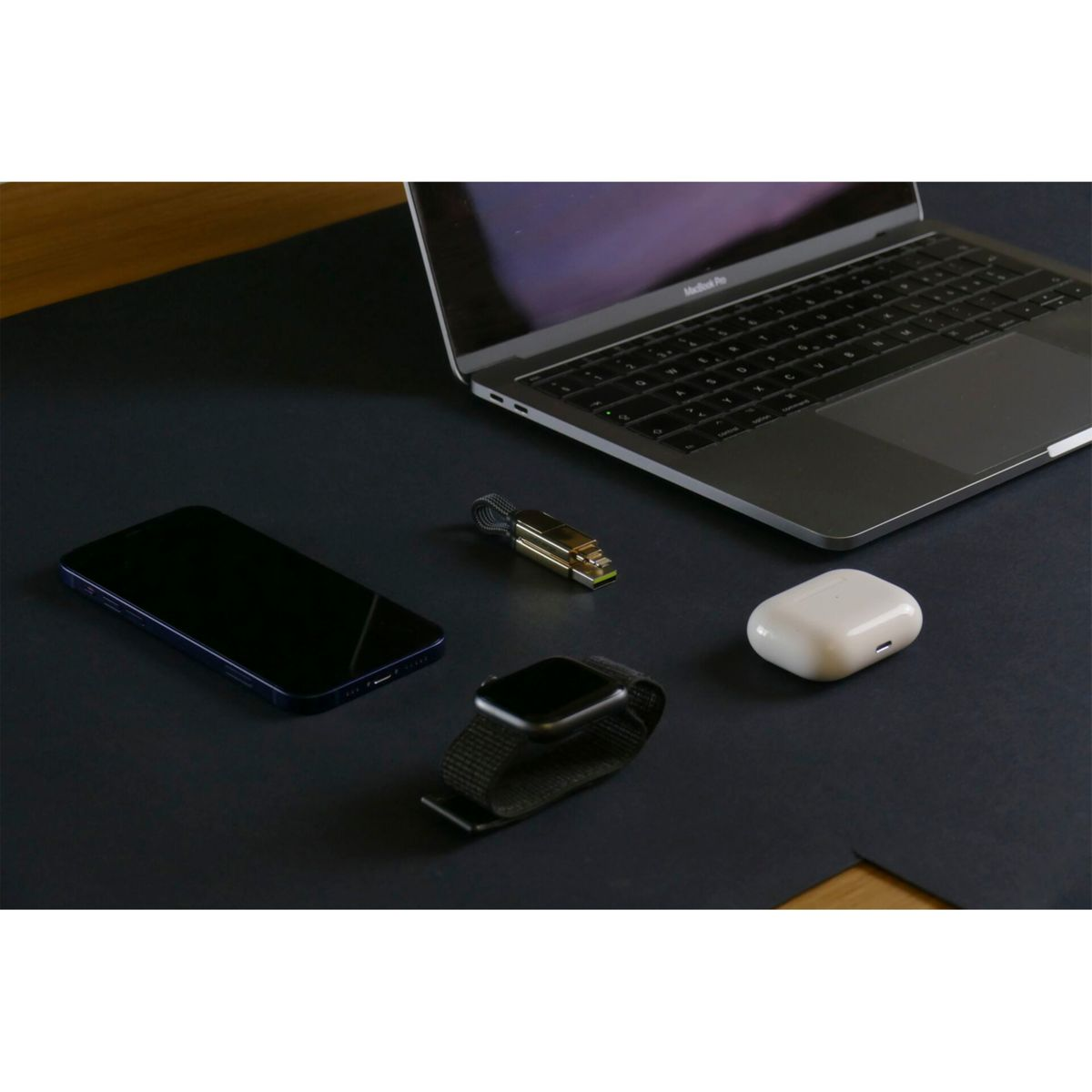 Ein Laptop, Handy, Smartwatch, USB-Stick und AirPods-Etui auf dunkler Oberfläche.