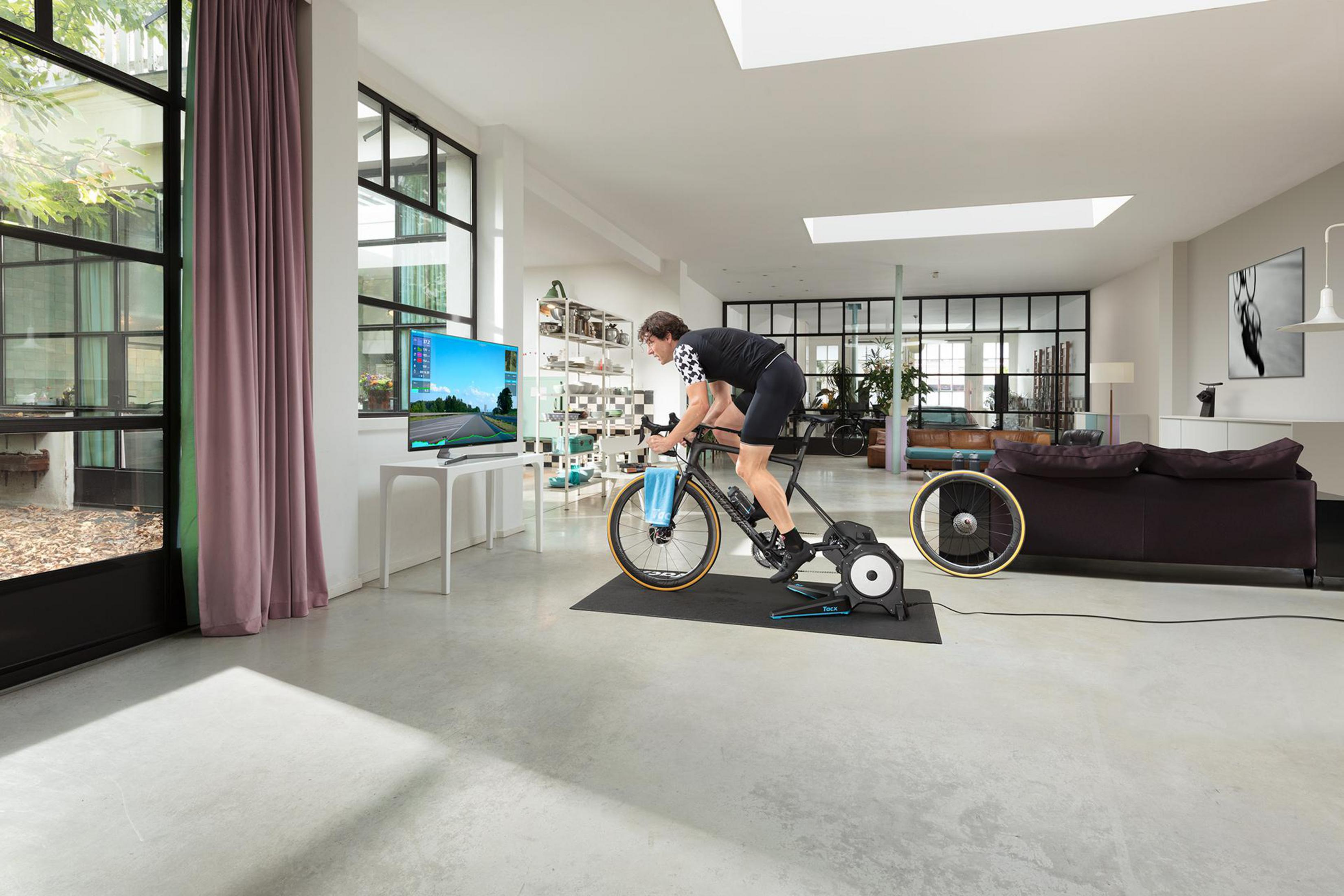 Uomo pedala su indoor trainer davanti a TV in soggiorno moderno.