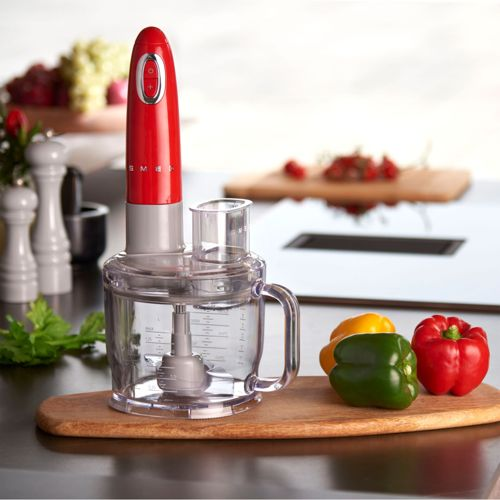 Procesador de alimentos Smeg rojo con bol transparente sobre tabla de madera, con pimientos.
