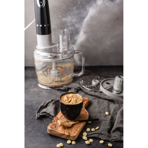 Un procesador de alimentos con un bol de mantequilla de frutos secos y chips de chocolate blanco cerca.